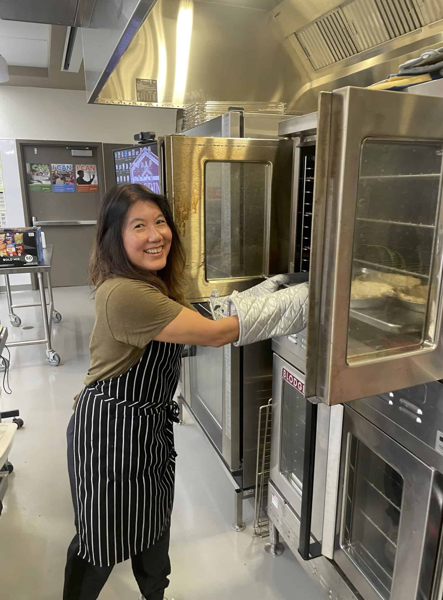 An Asian White House chef wearing an apron and oven mitts smiles while placing or removing a tray from a commercial oven in a kitchen.