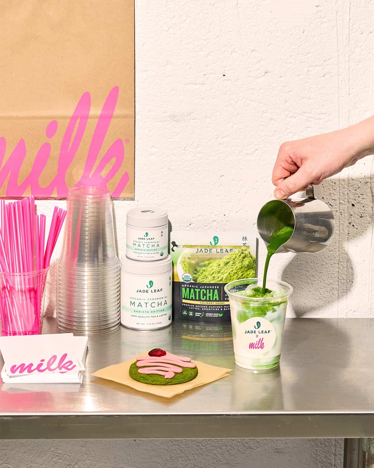 A hand pours matcha into a clear cup of milk beside containers of Jade Leaf Matcha, a bag of matcha powder, a pastry with pink frosting, stacked pink straws, plastic cups, and a pink “milk” sign on a metal counter.