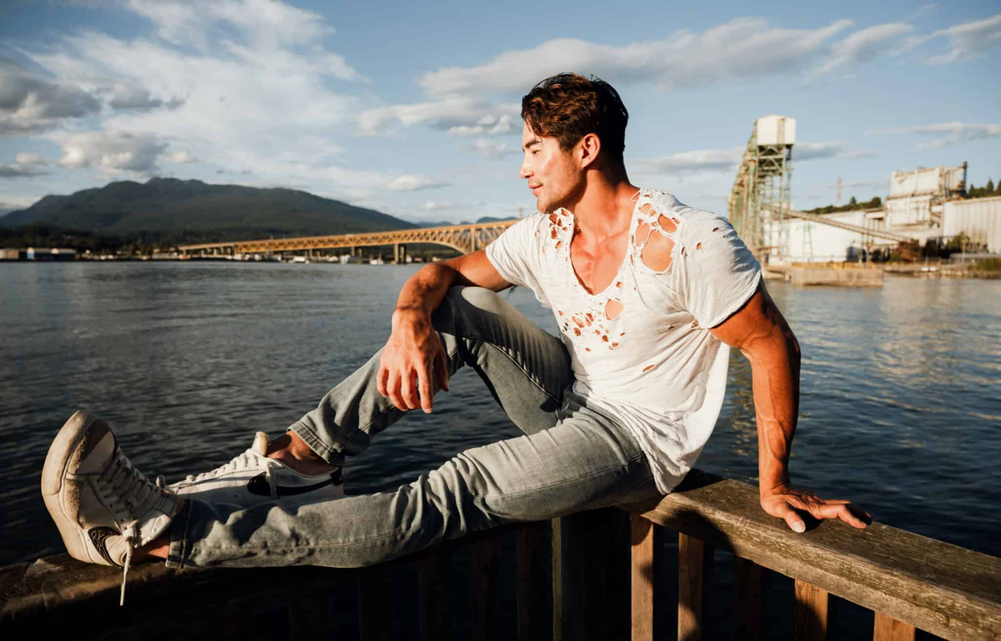 A young man in a ripped white t-shirt and jeans sits casually on a wooden railing by the water, with an industrial backdrop and mountains in the distance under a blue sky.