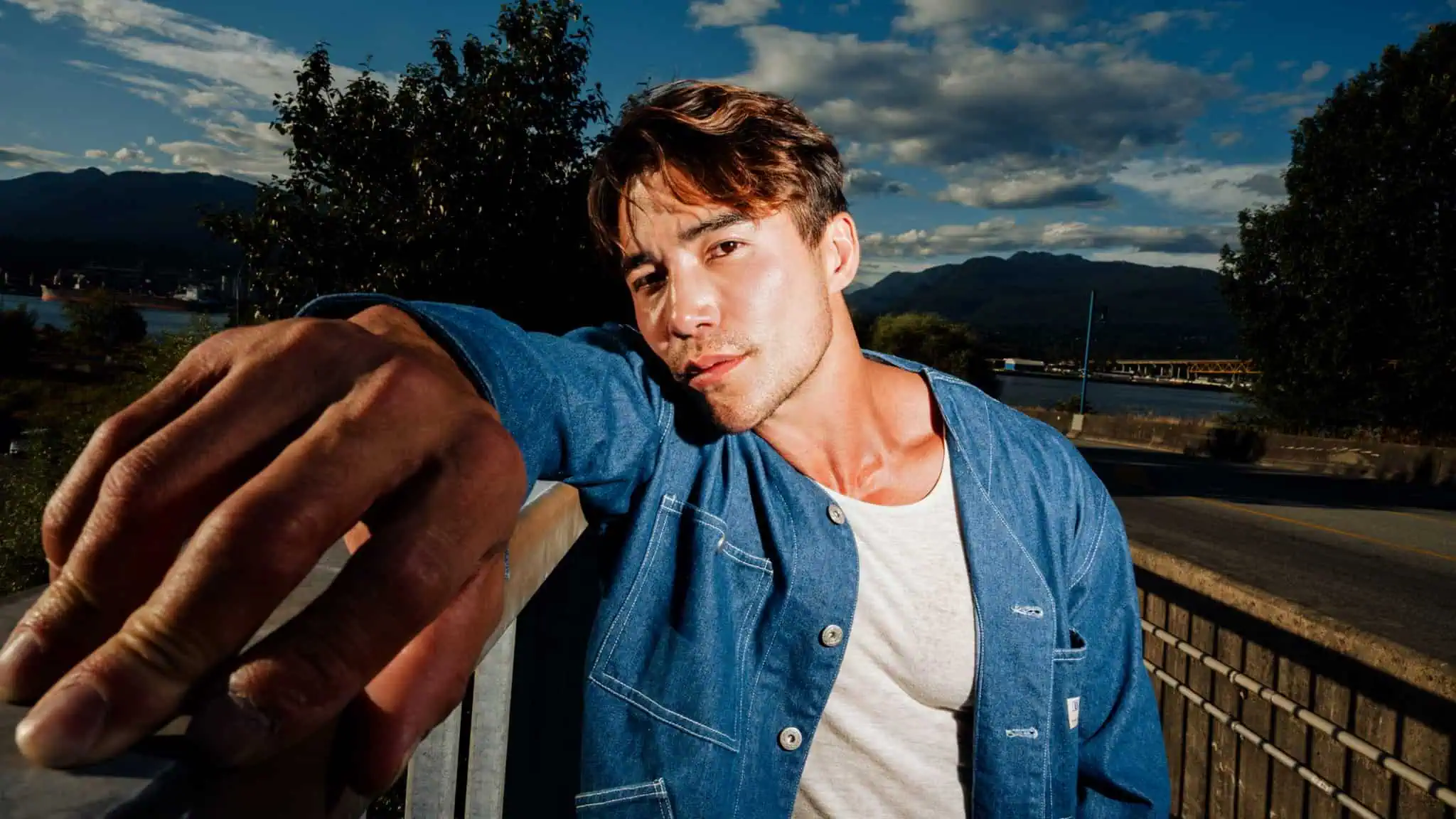 A man wearing a blue jacket and white shirt leans against a railing outdoors, extending his arm toward the camera. There are trees, mountains, and a partly cloudy sky in the background.