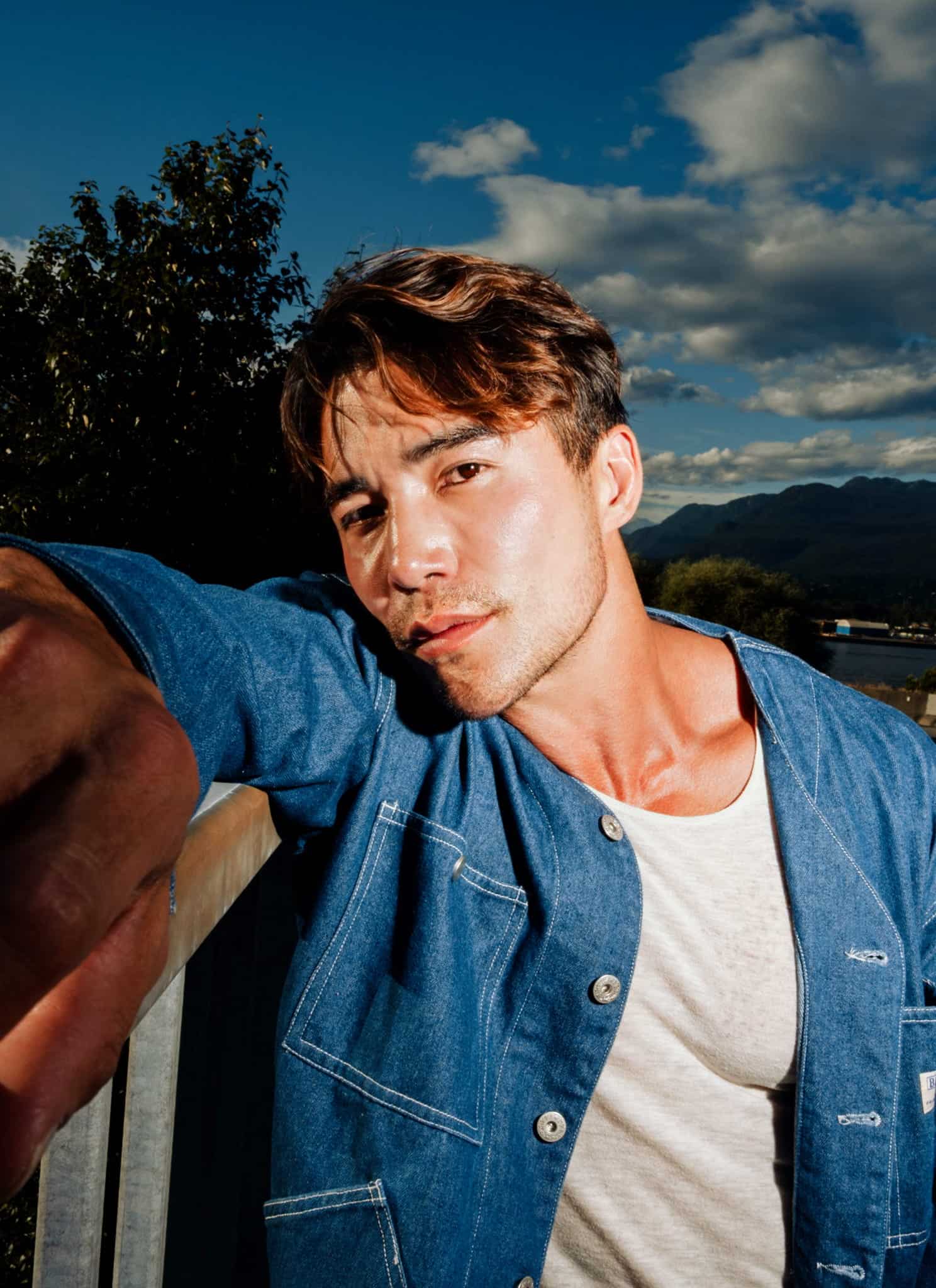 A man wearing a blue jacket and white shirt leans against a railing outdoors, extending his arm toward the camera. There are trees, mountains, and a partly cloudy sky in the background.