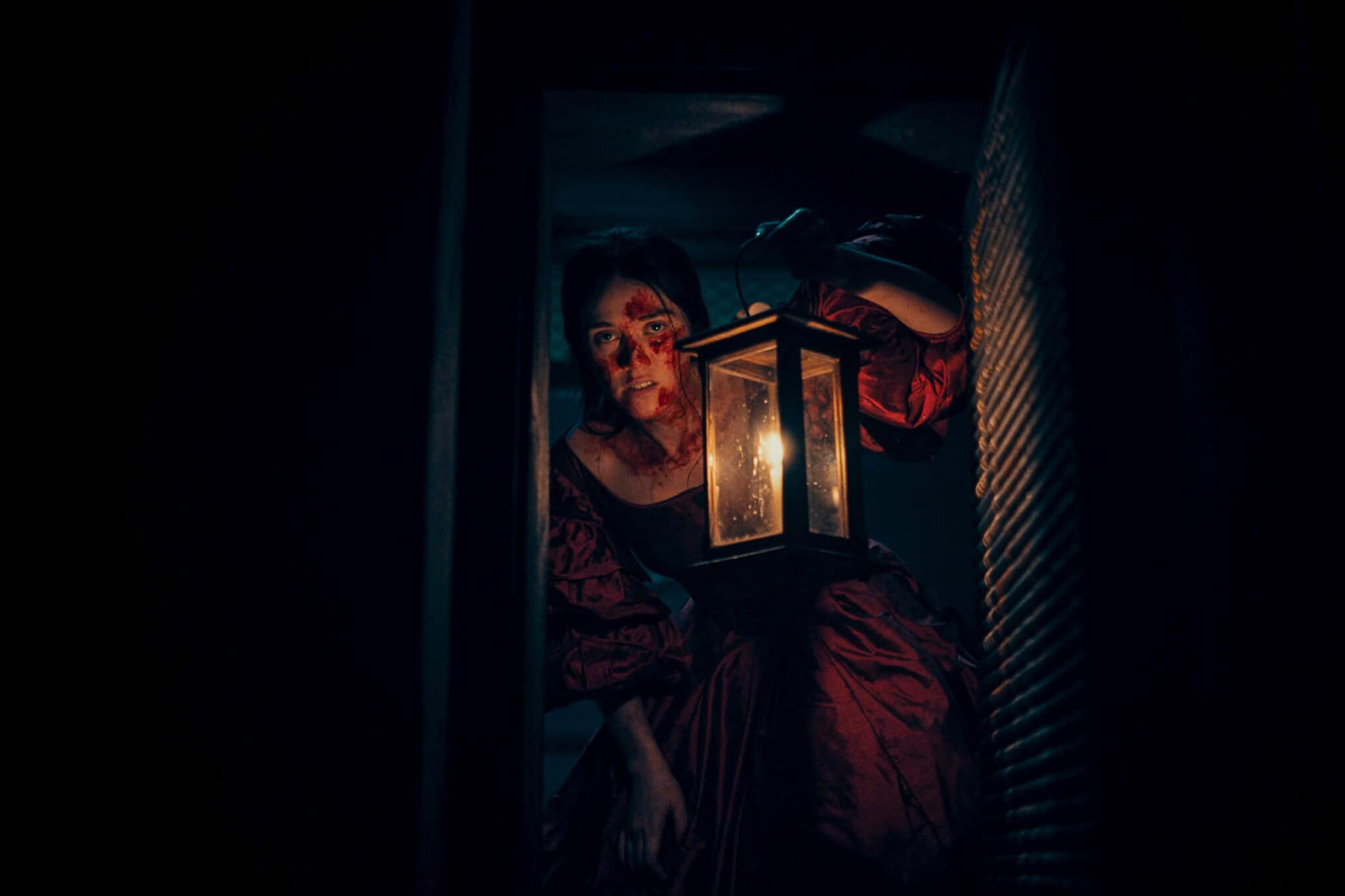 A woman in a dark, old-fashioned dress holds a lit lantern, her face streaked with blood, and peers through a doorway into the darkness, creating a tense, eerie atmosphere.