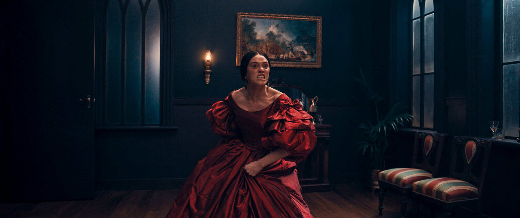 A woman in a dramatic red gown stands in a dimly lit, elegant room, clenching her fist and snarling angrily. Dark walls, a painting, arched windows, and striped chairs complete the moody, historical setting.
