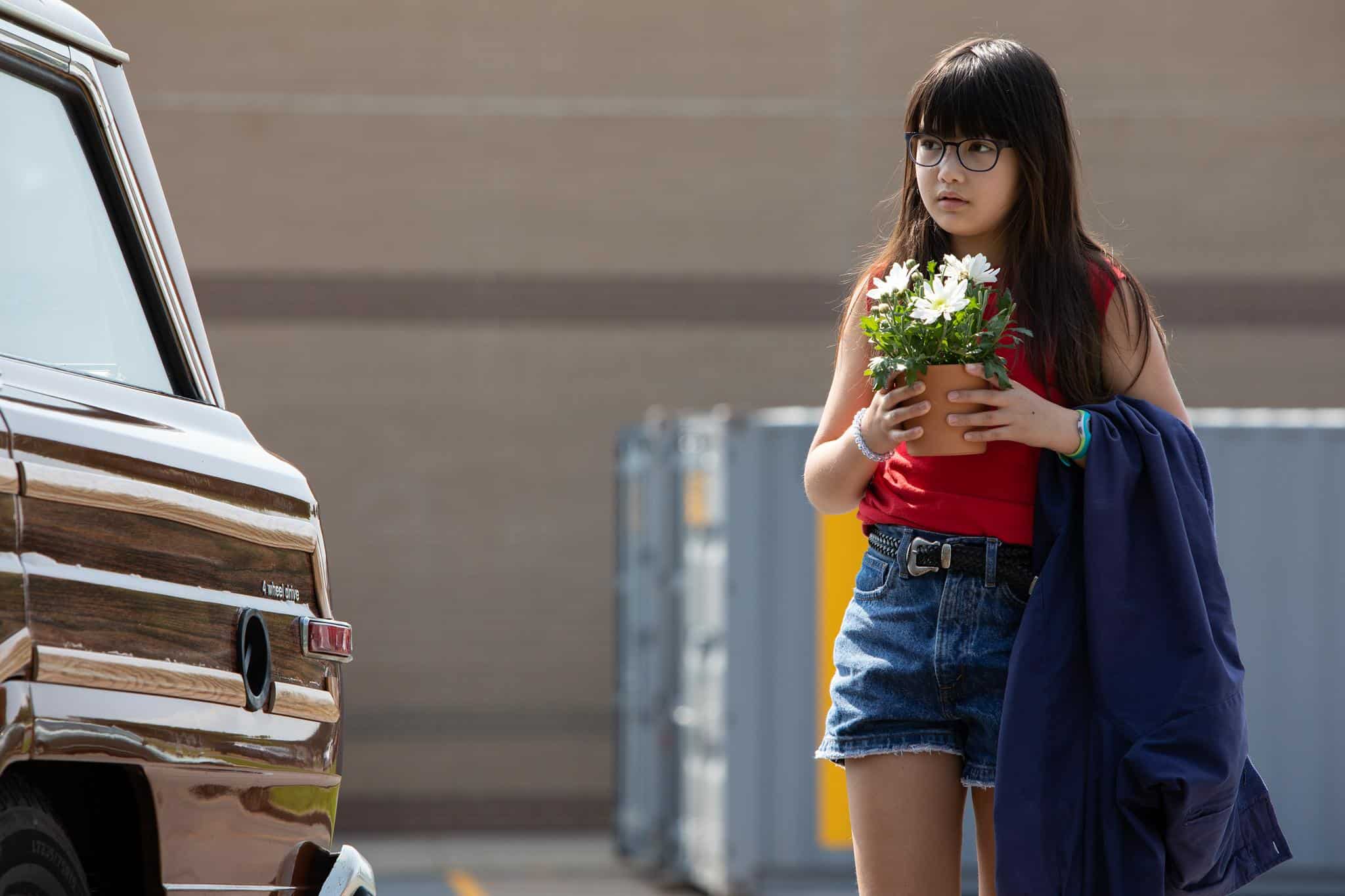 A girl with long dark hair and glasses, wearing a red tank top and denim shorts, holds a potted plant and a blue jacket while standing next to a parked vehicle in an outdoor setting.
