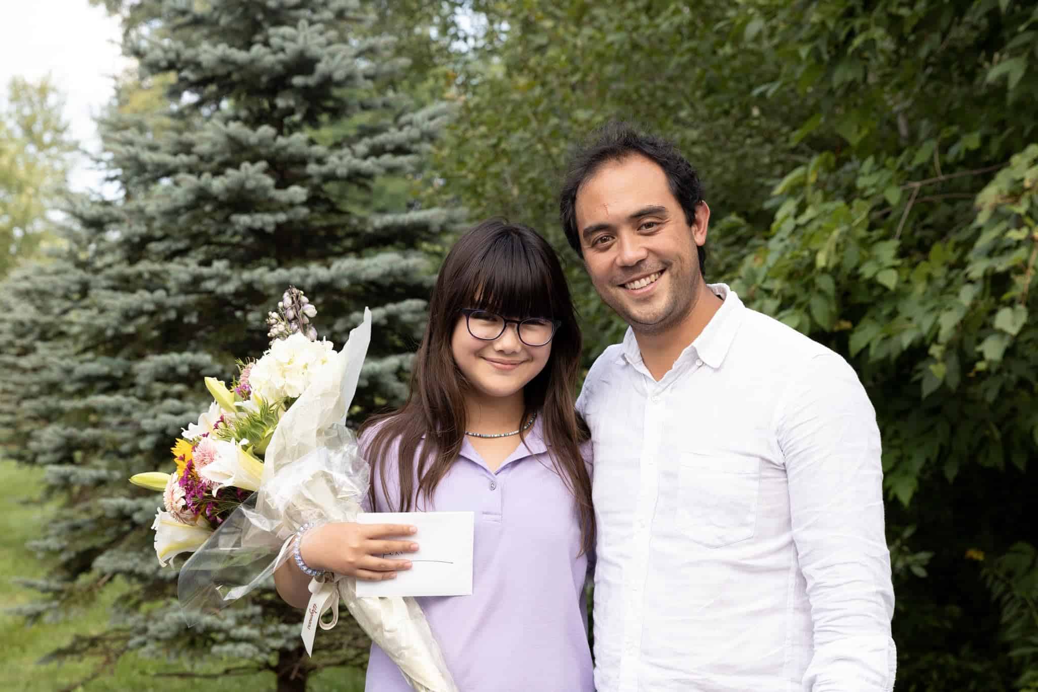 A young girl holding a bouquet of flowers and an envelope stands next to a smiling man in a white shirt, with trees and greenery in the background.