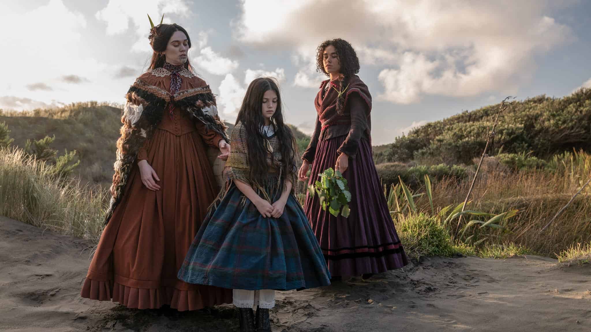 Three women in Victorian-era dresses stand outdoors on sandy terrain, surrounded by grass and bushes. One woman holds leaves, another has a feathered cape, and a young girl in a blue plaid dress stands between them, looking down.