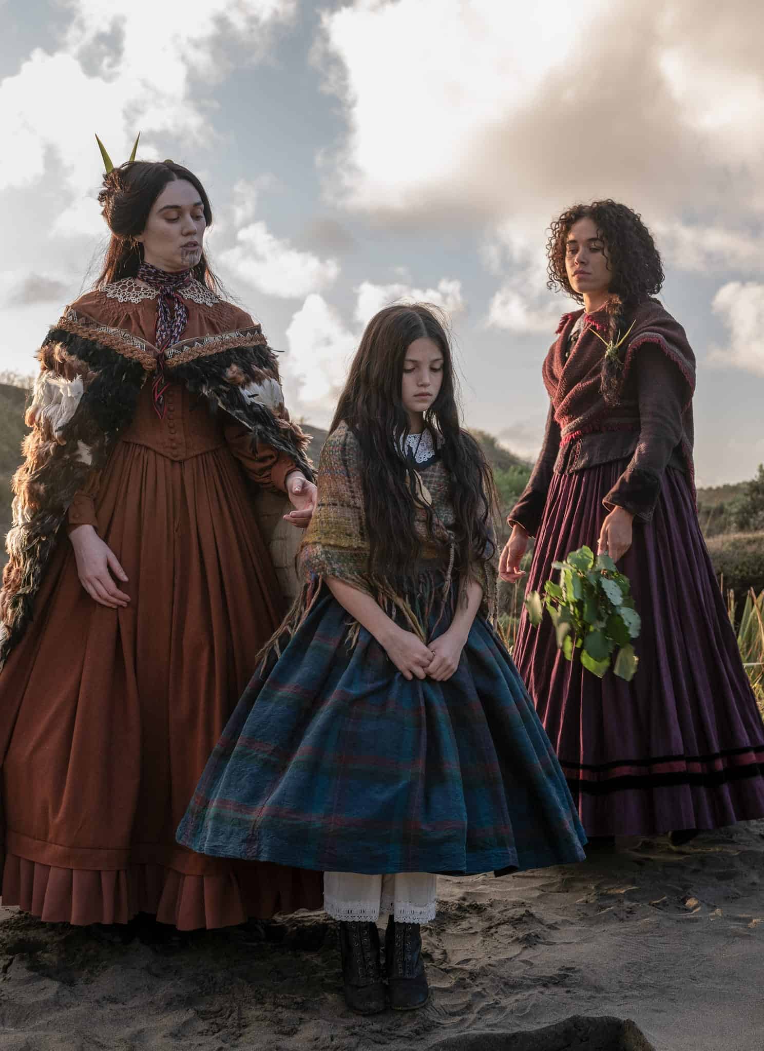 Three women in Victorian-era dresses stand outdoors on sandy terrain, surrounded by grass and bushes. One woman holds leaves, another has a feathered cape, and a young girl in a blue plaid dress stands between them, looking down.
