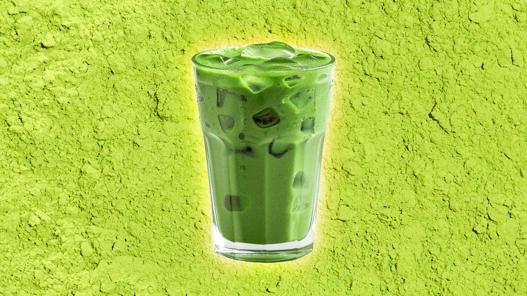 A tall glass of iced matcha latte with ice cubes, set against a bright green background that resembles powdered matcha.