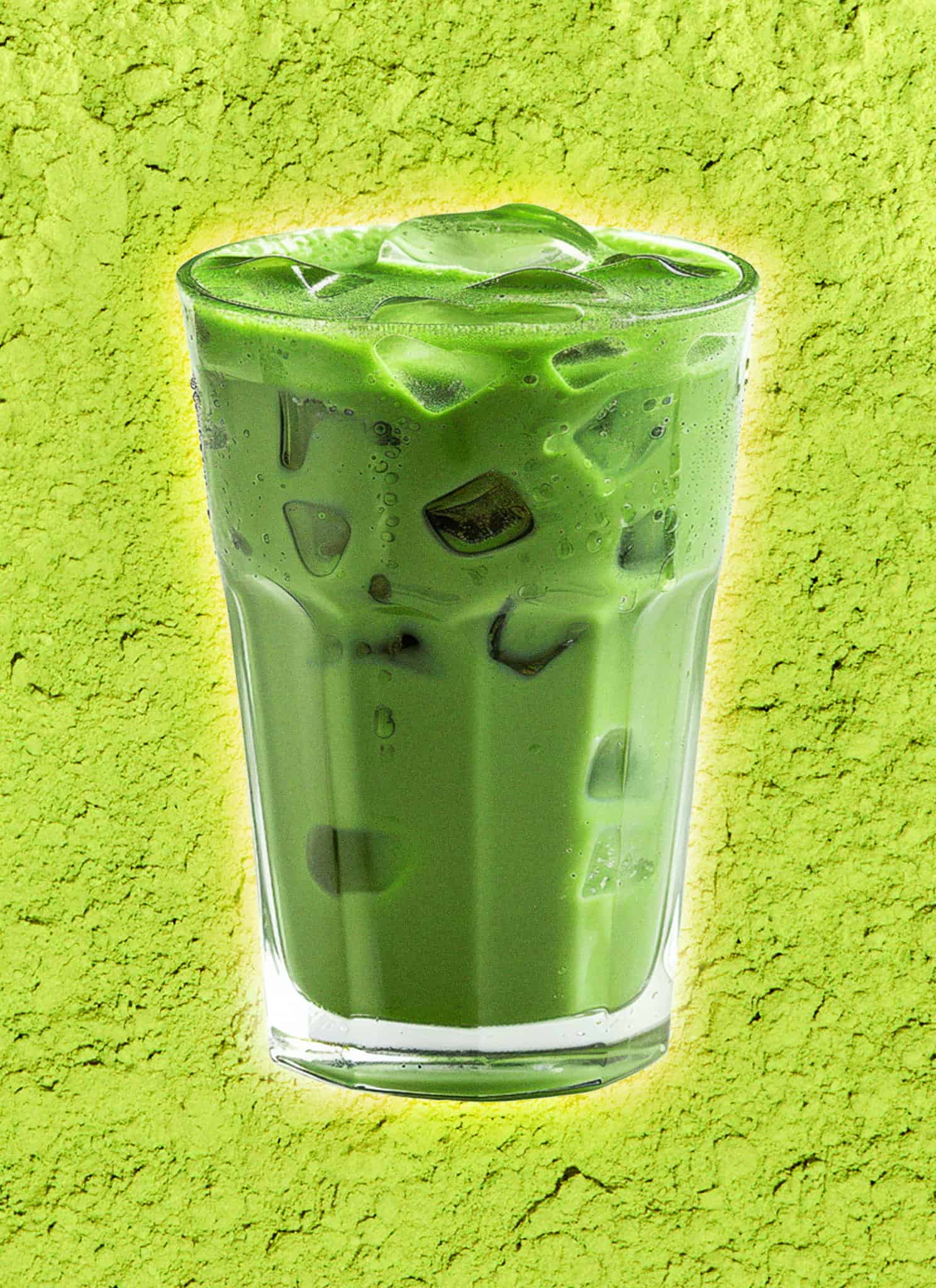 A tall glass of iced matcha latte with ice cubes, set against a bright green background that resembles powdered matcha.