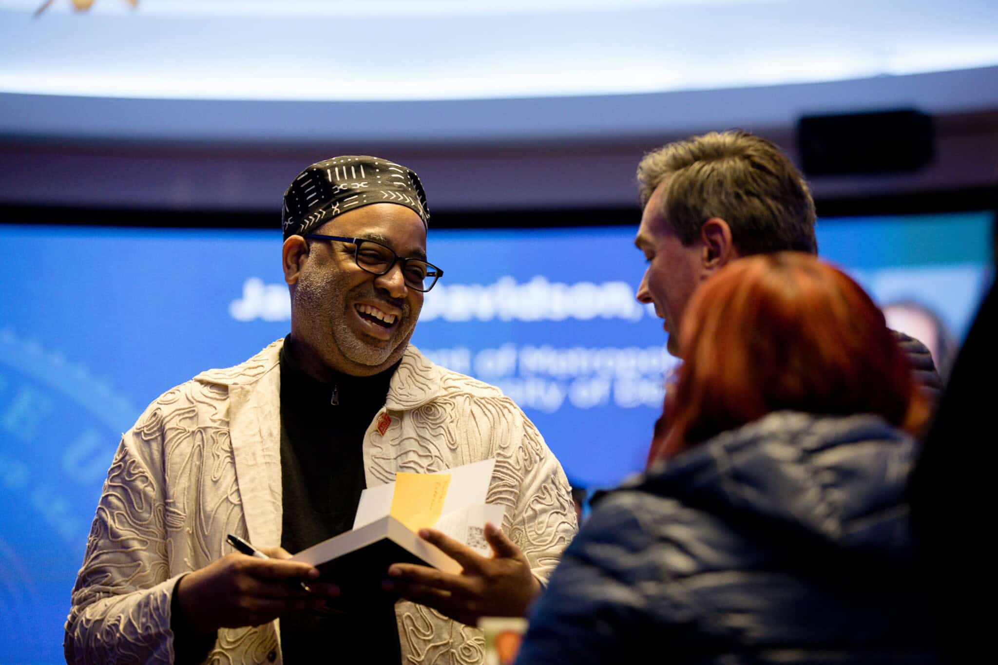 A smiling man in glasses and a patterned cap holds a book and pen while talking with two people in a warmly lit indoor setting, with a blurred presentation screen about Asian White House chefs in the background.
