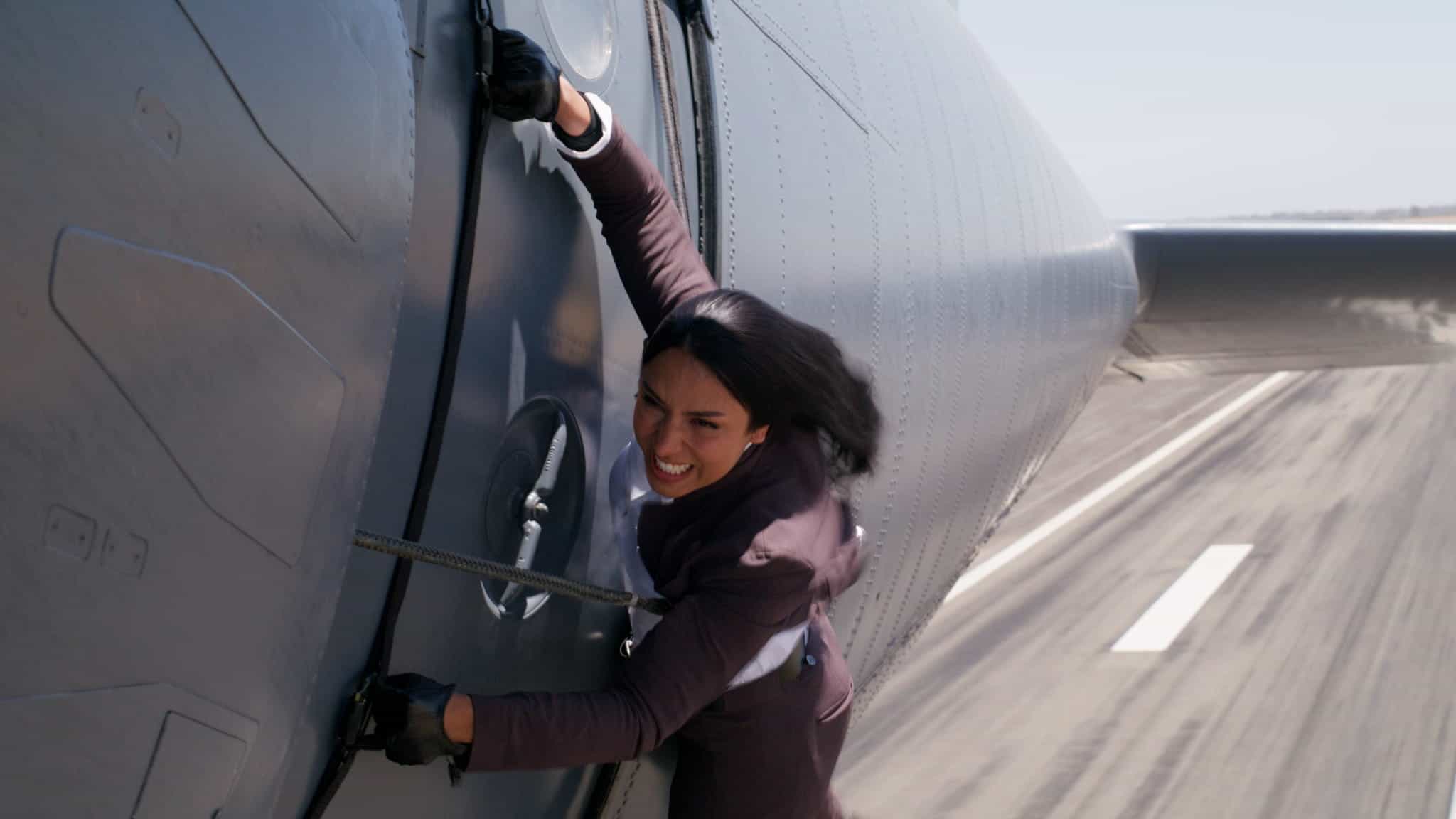 A woman in a suit clings to the side of a flying airplane, gripping a handle and looking distressed, with blurred runway visible below.