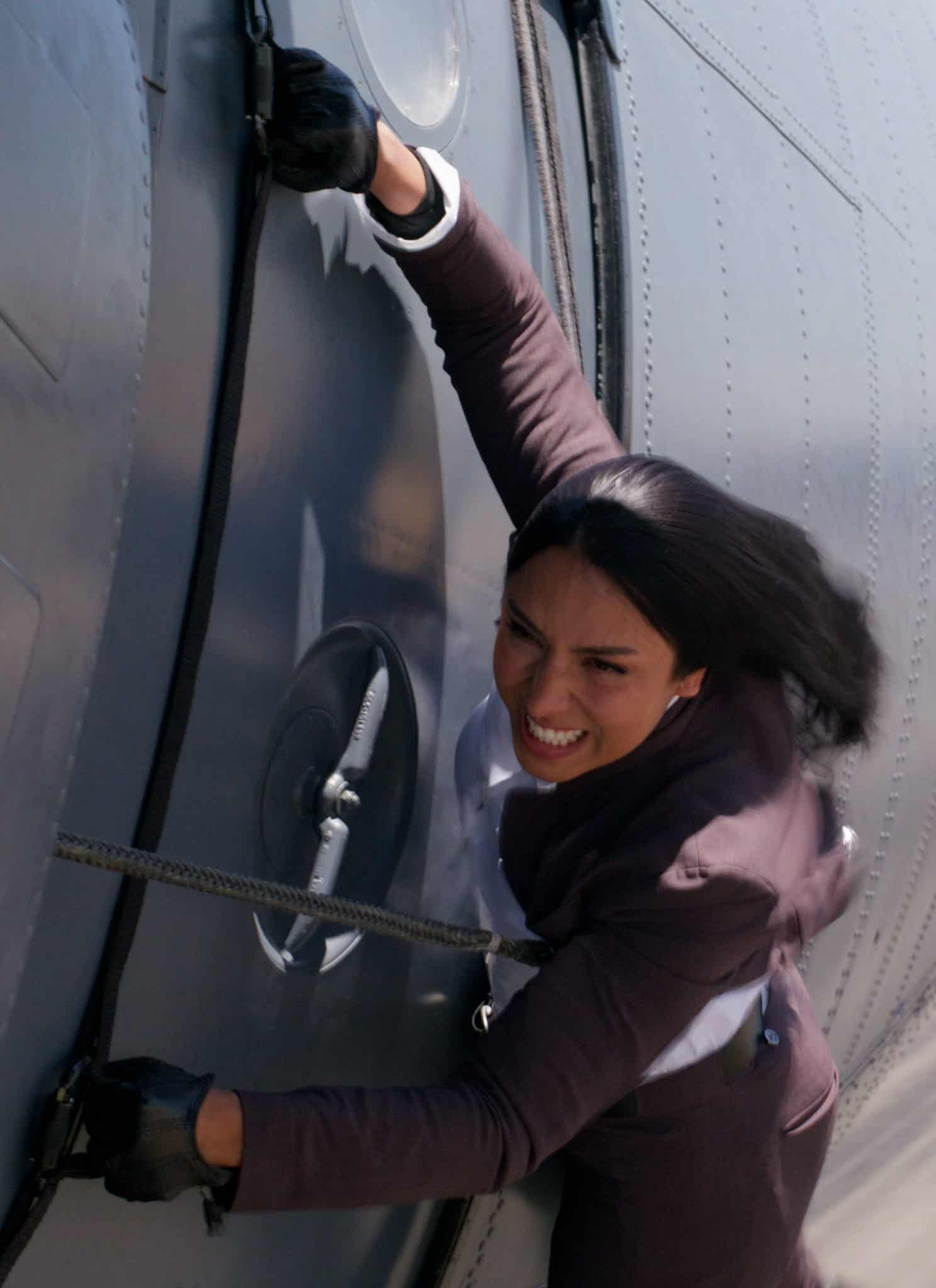 A woman in a suit clings to the side of a flying airplane, gripping a handle and looking distressed, with blurred runway visible below.