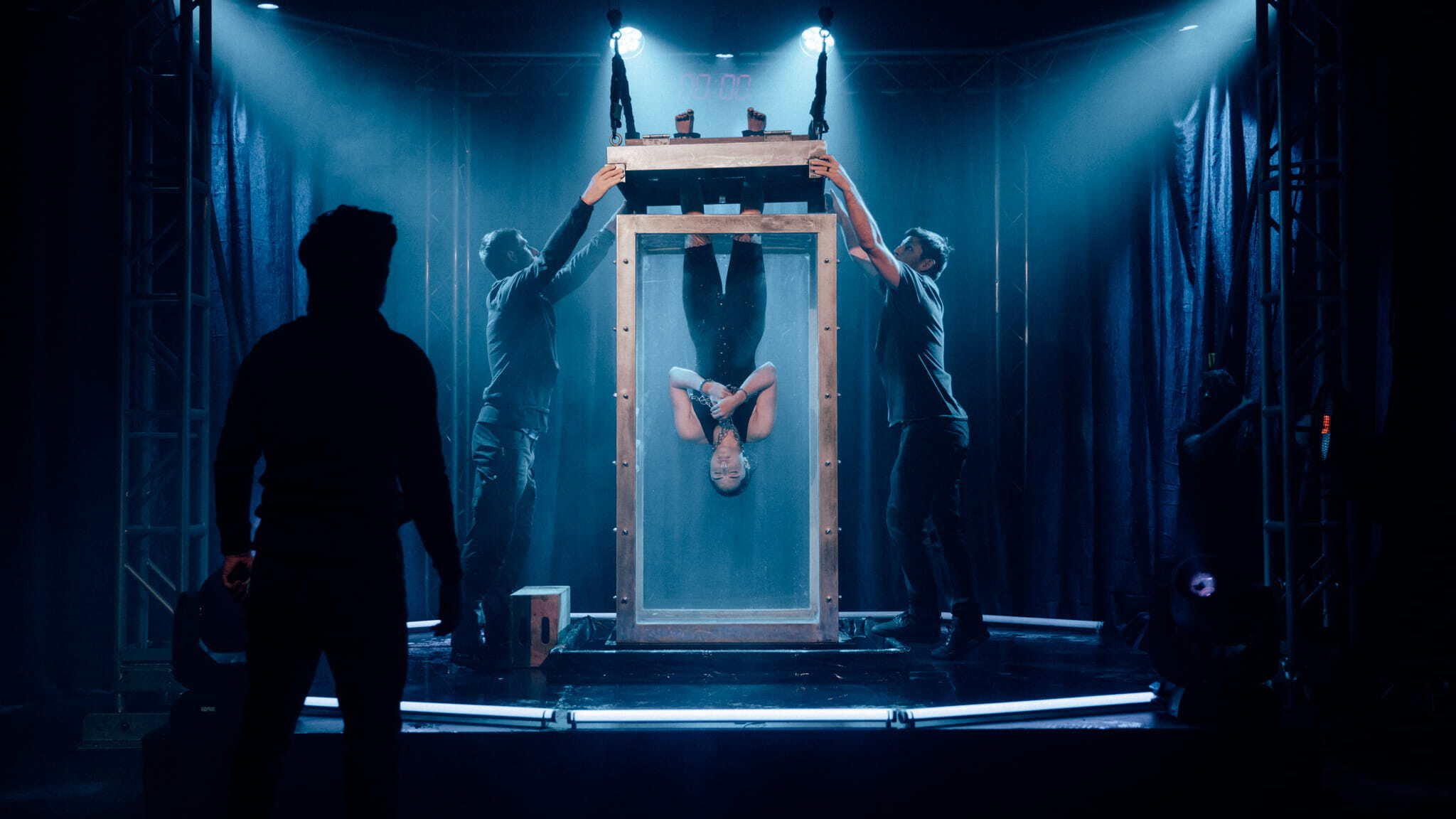 A person is suspended upside down and submerged in a glass water tank on stage, while two assistants hold the tank’s lid. Another figure stands in shadow, watching amid dramatic blue stage lighting.