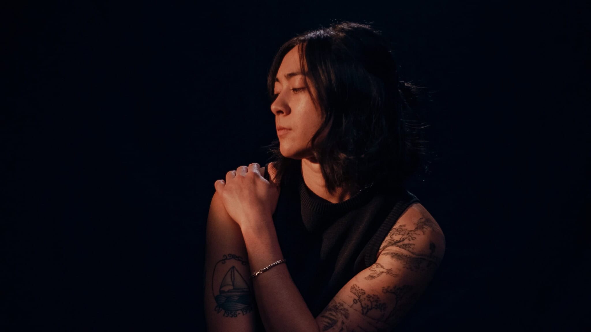 A person with shoulder-length dark hair and tattoos on their arms sits against a dark background, looking down and to the side with one hand resting on their opposite shoulder—soft, moody lighting.