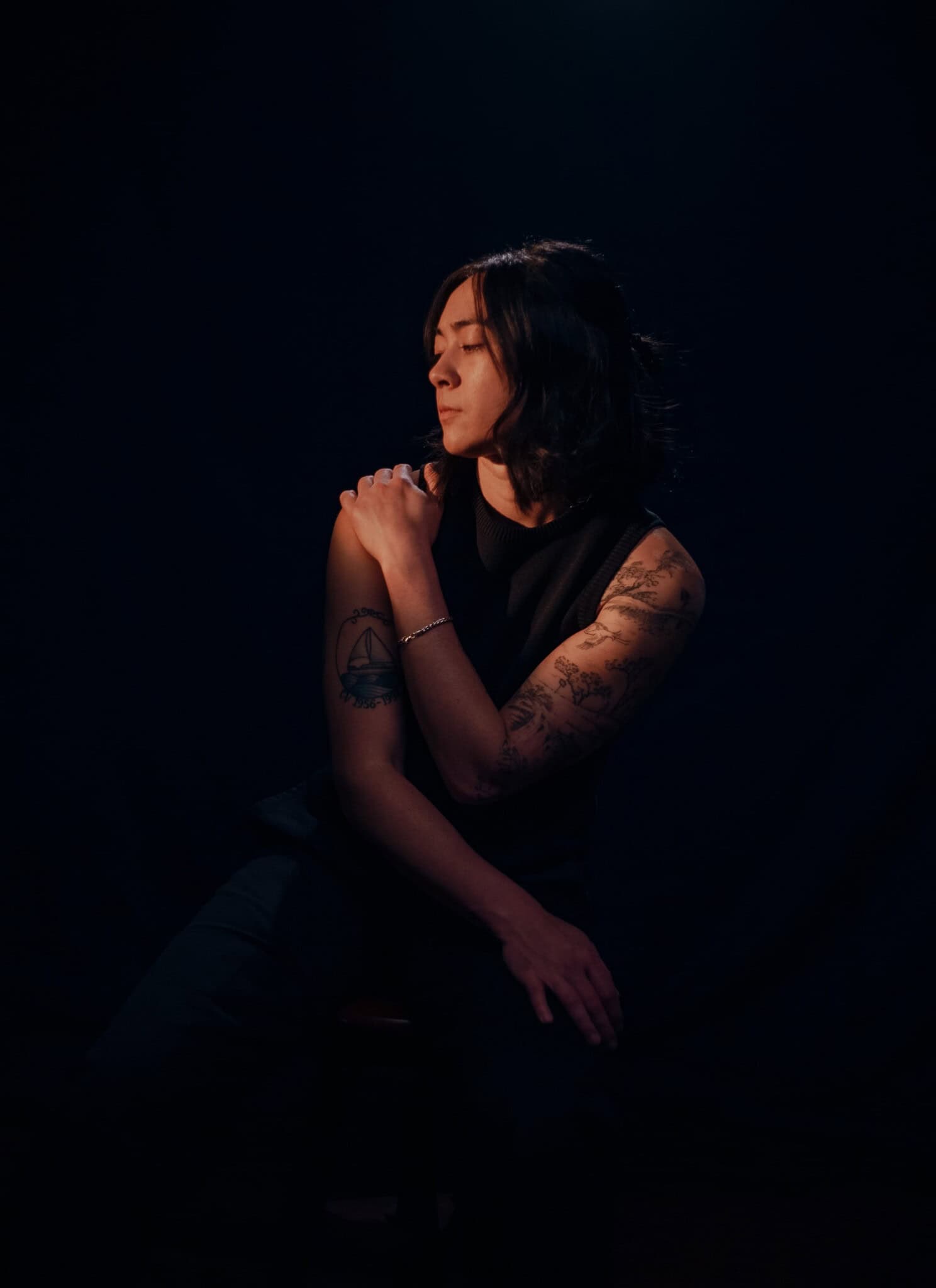 A person with shoulder-length dark hair and tattoos on their arms sits against a dark background, looking down and to the side with one hand resting on their opposite shoulder—soft, moody lighting.