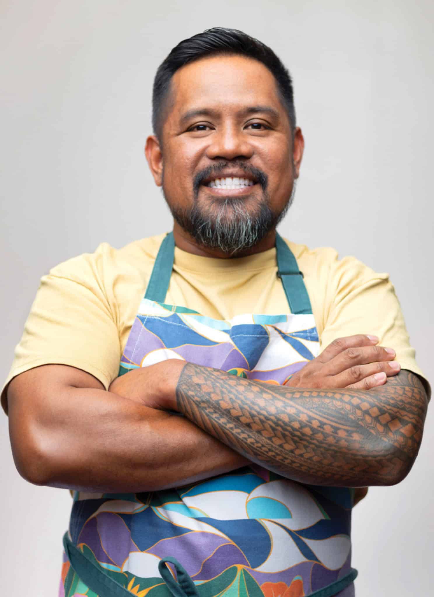 A man with short black hair and a beard smiles warmly at the camera. He is wearing a light yellow shirt and a green apron. The background is plain and light-colored.