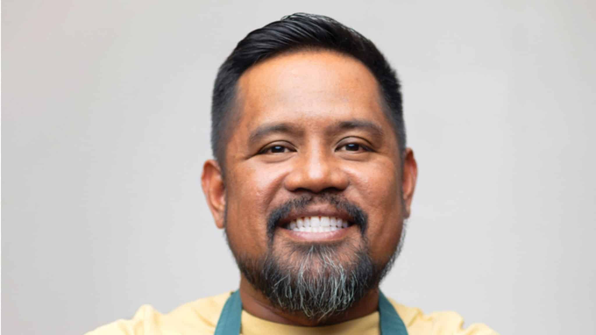 A man with short black hair and a beard smiles warmly at the camera. He is wearing a light yellow shirt and a green apron. The background is plain and light-colored.