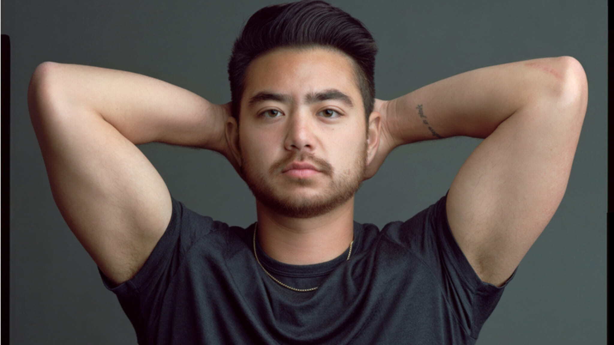 A person with short dark hair and a trimmed beard poses with their hands resting behind their head, wearing a dark t-shirt and a thin gold necklace against a plain gray background.