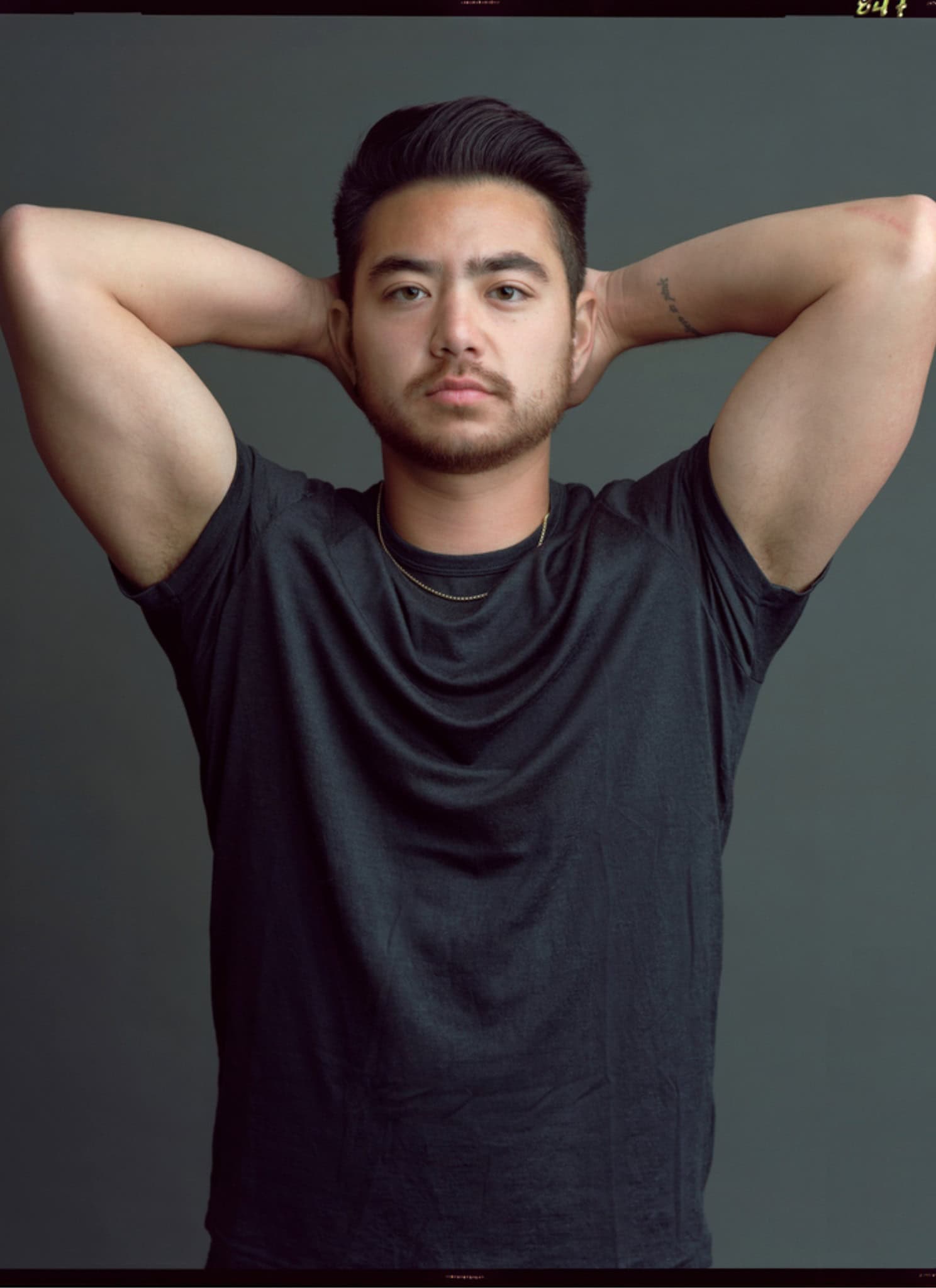 A person with short dark hair and a trimmed beard poses with their hands resting behind their head, wearing a dark t-shirt and a thin gold necklace against a plain gray background.