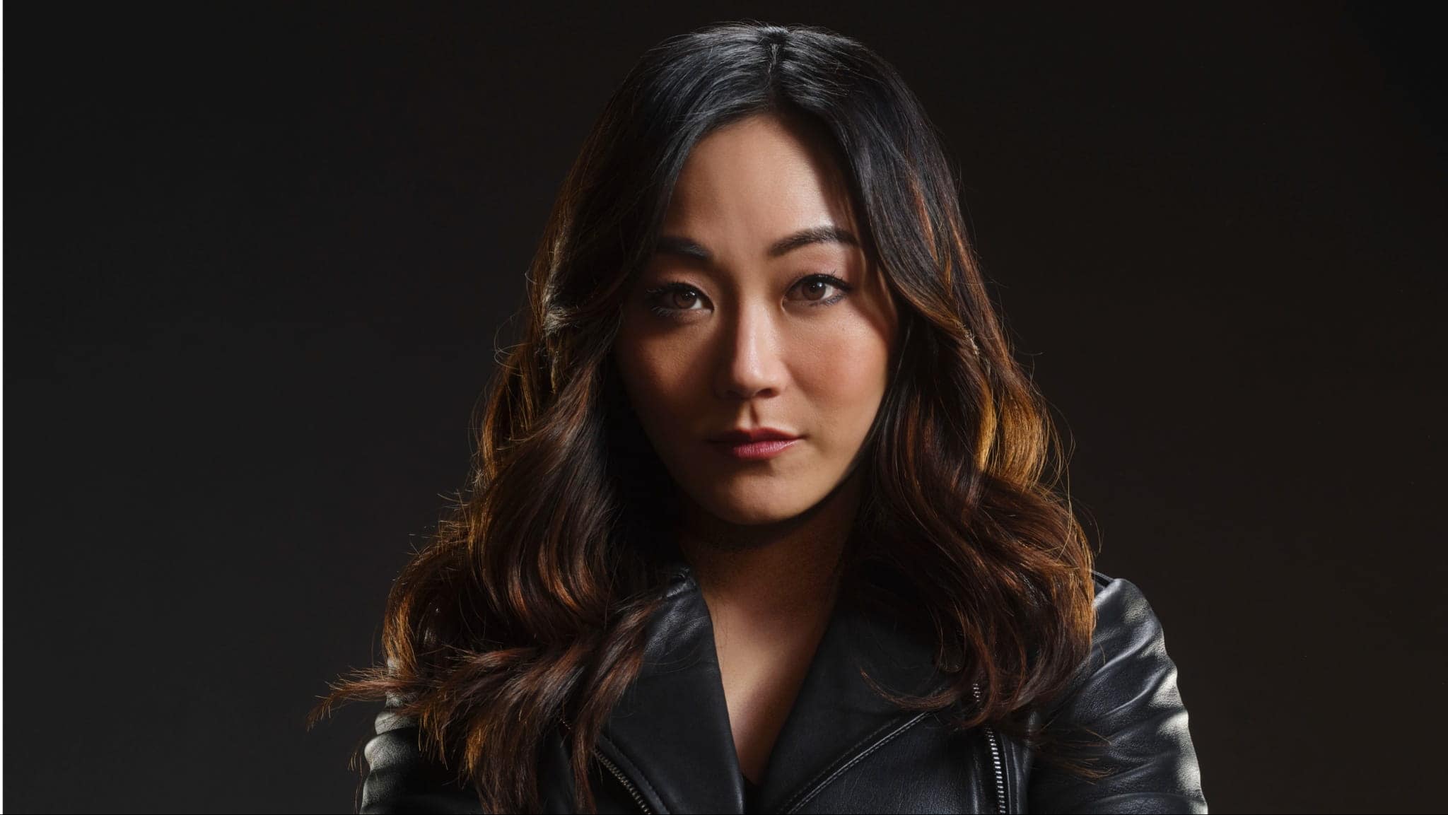 A woman with long, wavy dark hair and a serious expression, resembling Karen Fukuhara, wearing a black leather jacket, posed against a dark background.
