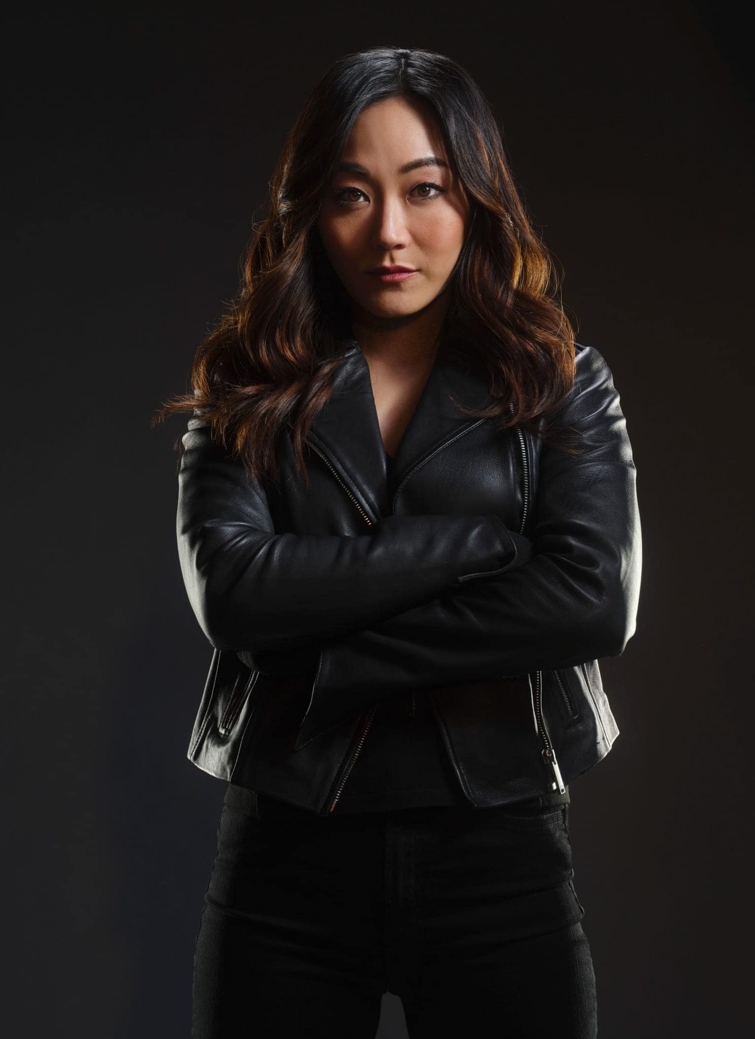 A woman with long, wavy dark hair and a serious expression, resembling Karen Fukuhara, wearing a black leather jacket, posed against a dark background.