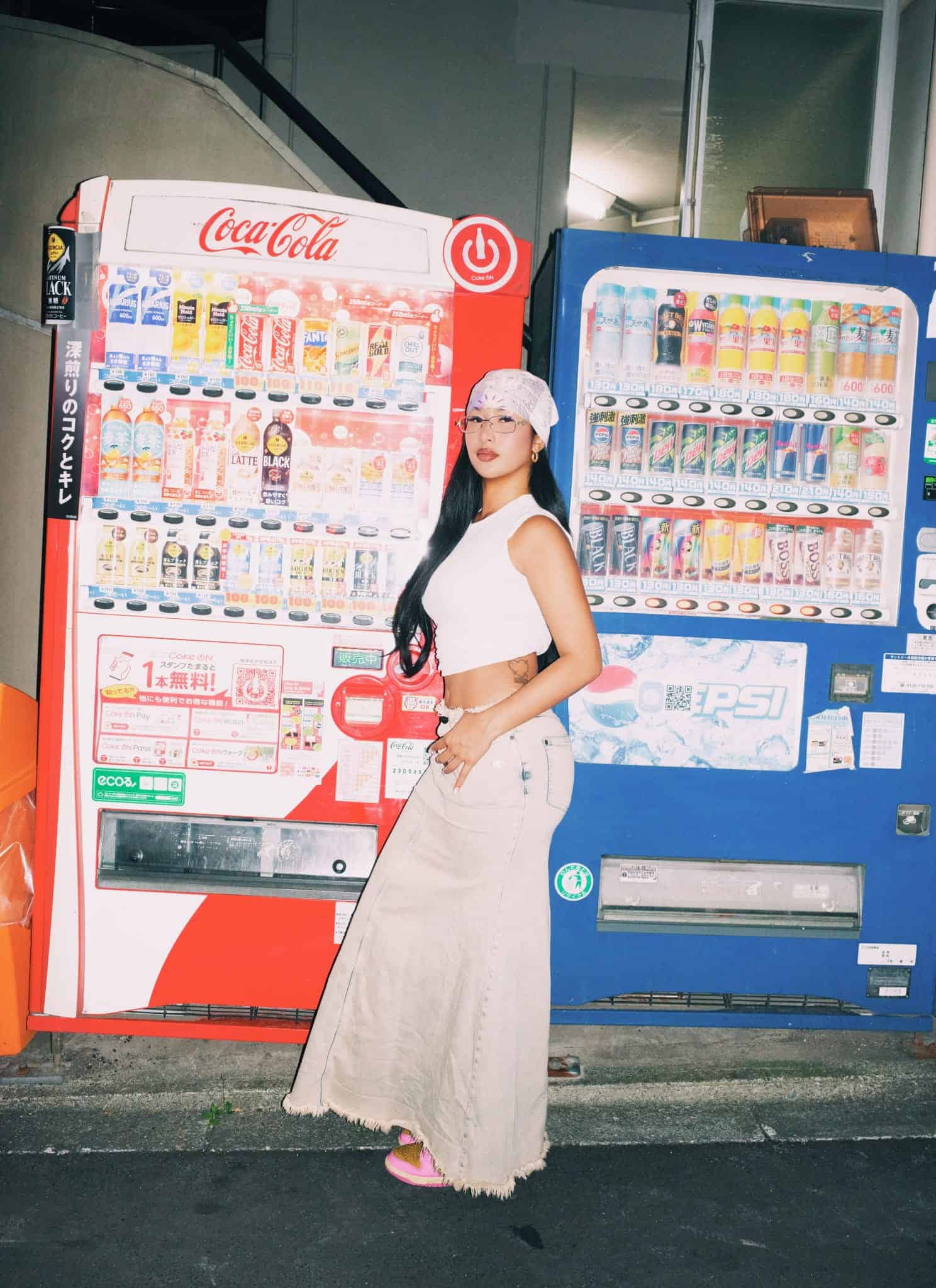 A person wearing a white sleeveless top, beige pants, a patterned bandana, and glasses stands in front of colorful Softest Hard vending machines filled with drinks, channeling stylish LE SSERAFIM vibes.