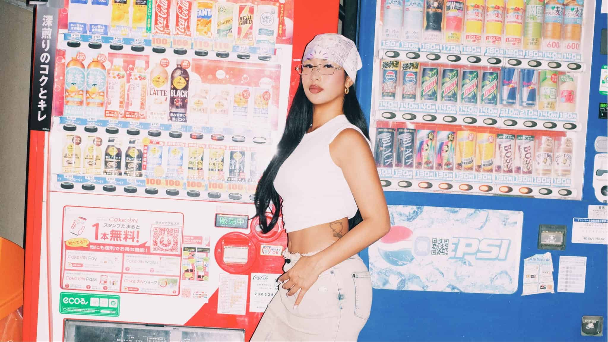 A person wearing a white sleeveless top, beige pants, a patterned bandana, and glasses stands in front of colorful Softest Hard vending machines filled with drinks, channeling stylish LE SSERAFIM vibes.