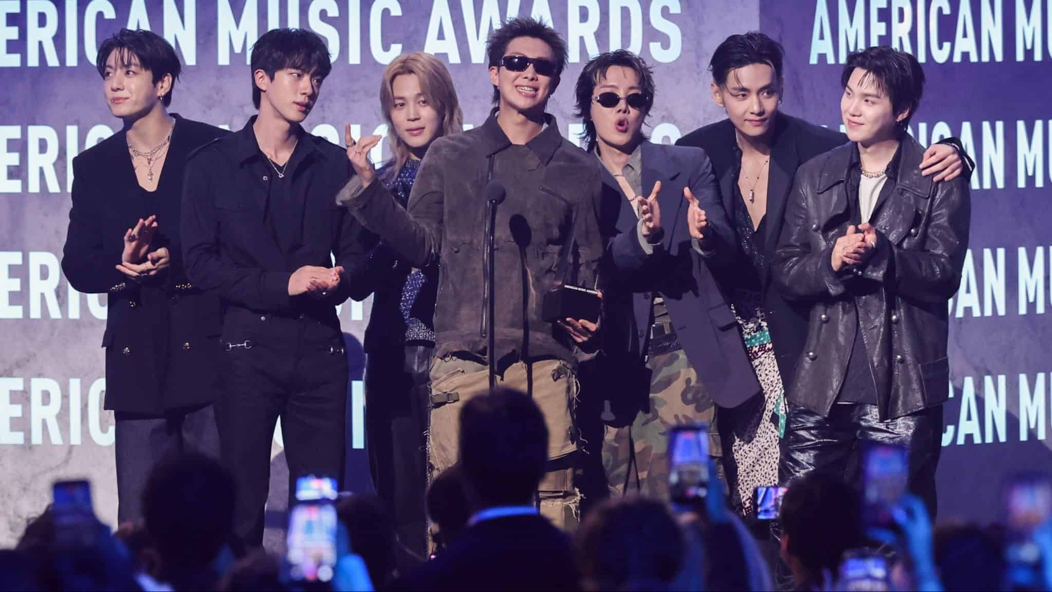 Seven men stand together on stage in front of a microphone at an awards show, smiling and gesturing towards the audience, with AMERICAN MUSIC AWARDS visible on the background screen behind them.