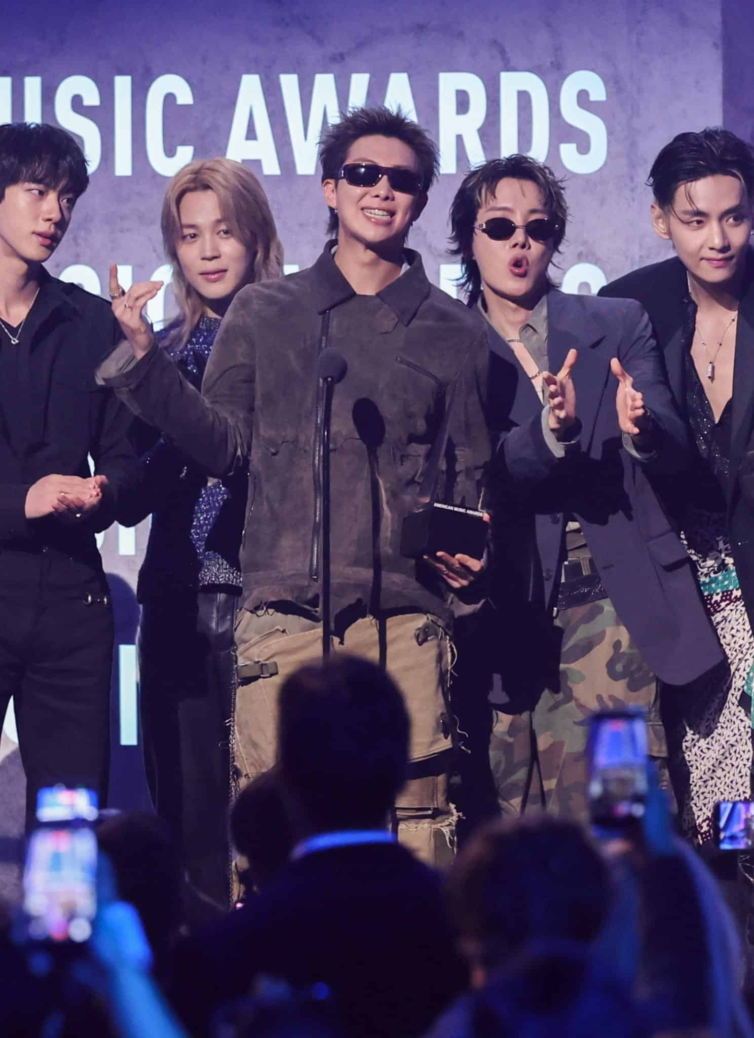Seven men stand together on stage in front of a microphone at an awards show, smiling and gesturing towards the audience, with AMERICAN MUSIC AWARDS visible on the background screen behind them.