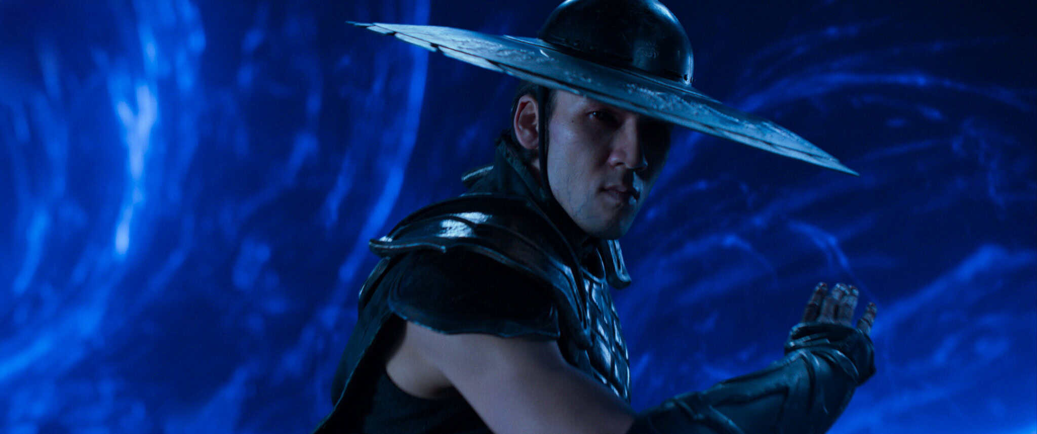 A man in dark armor poses for combat, wearing a wide, bladed hat in a dramatic blue-lit setting with swirling energy patterns in the background.