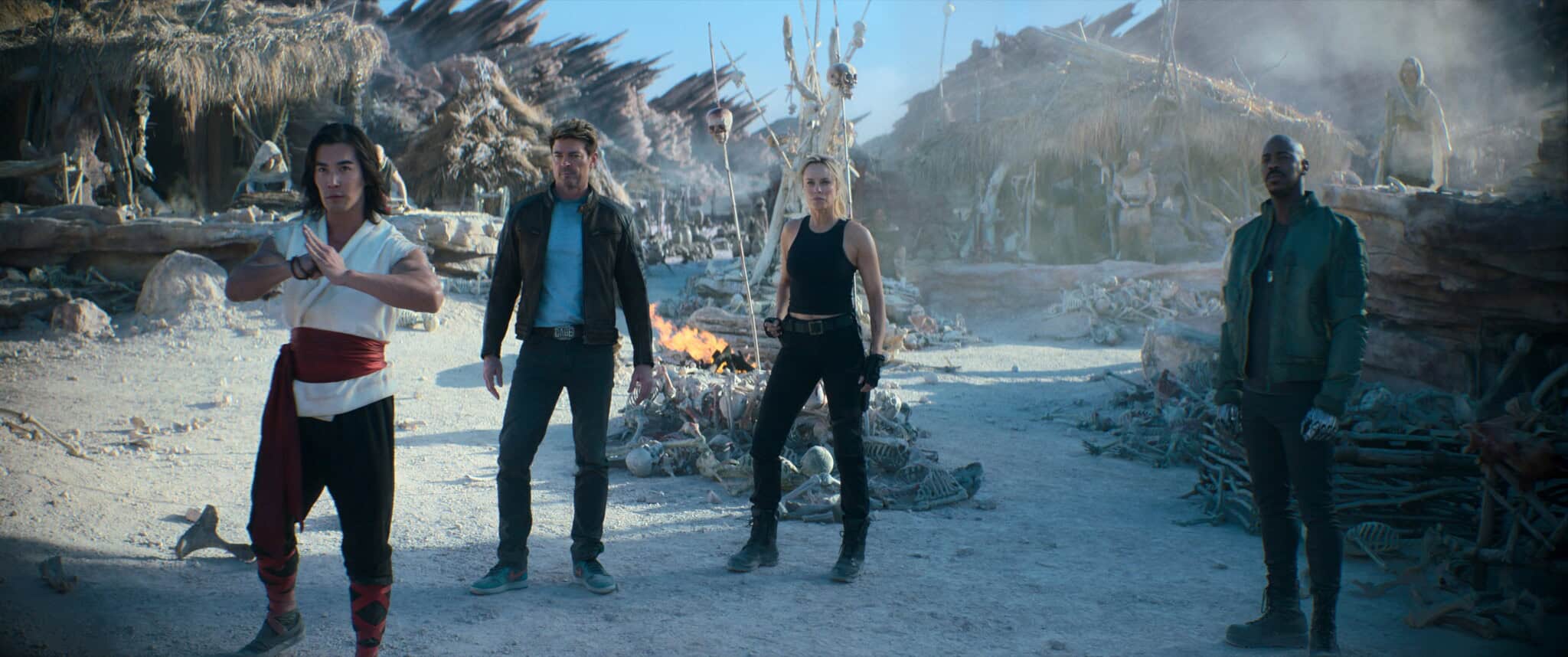 Four people stand in a barren, rocky landscape with skeletal remains and debris; one is in a martial arts stance, while the others stand alert, ready for action. A fire burns in the background among destroyed huts.