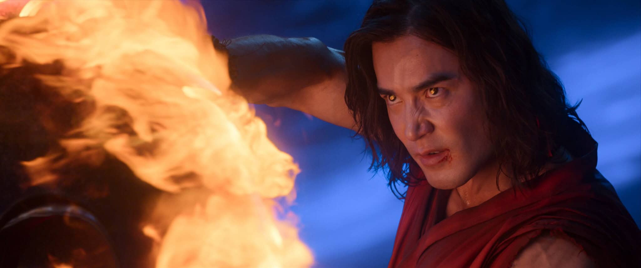 A man with long dark hair and a serious expression holds his arm up to a large burst of flames, with firelight illuminating his face. He wears a red outfit and stands against a blue-toned background.