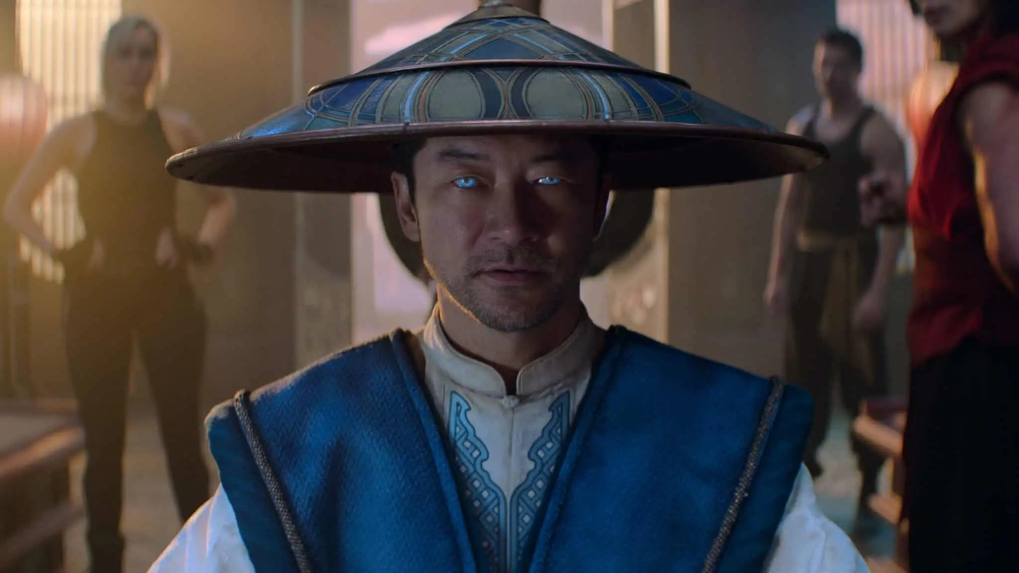 A man wearing a wide-brimmed hat and blue robe with glowing blue eyes stands indoors, facing the camera, with two people blurred in the background.