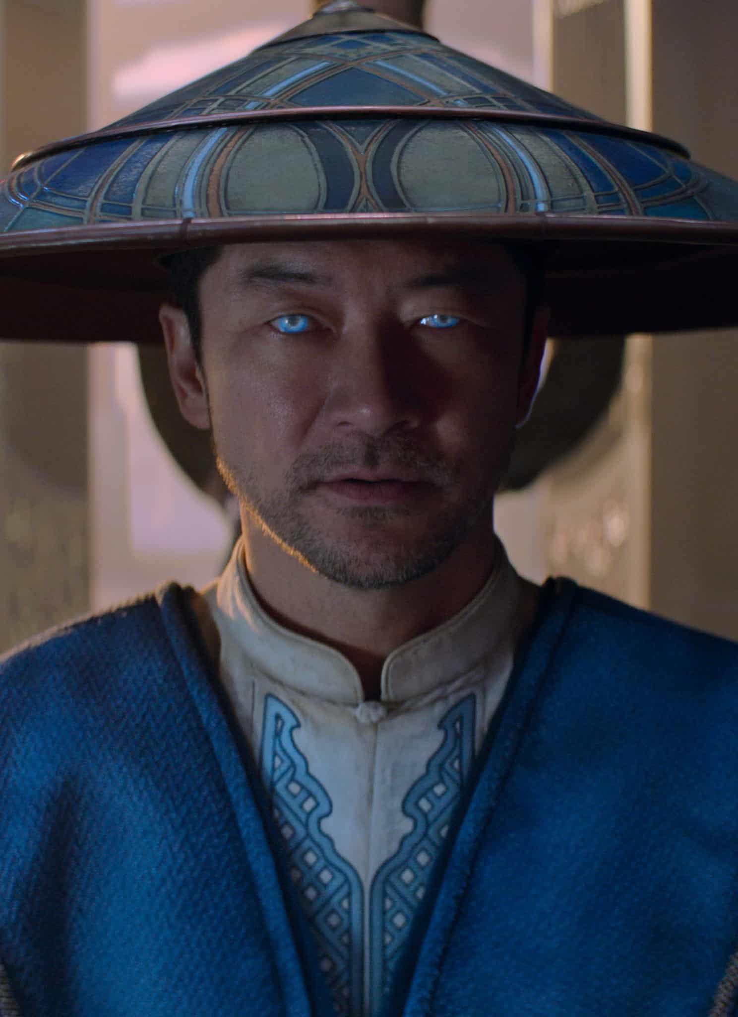 A man wearing a wide-brimmed hat and blue robe with glowing blue eyes stands indoors, facing the camera, with two people blurred in the background.