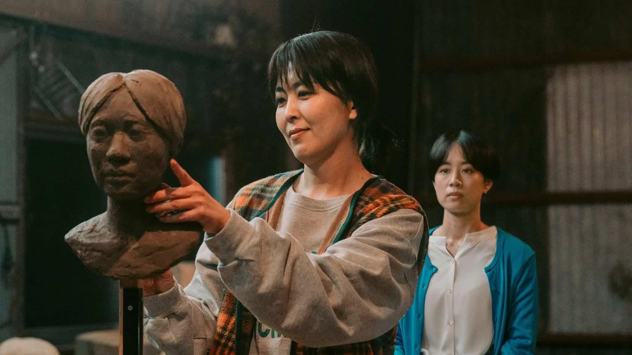 A woman in a plaid vest sculpts a clay bust, focusing intently, while another woman in a blue cardigan stands in the background watching her in a dimly lit studio.
