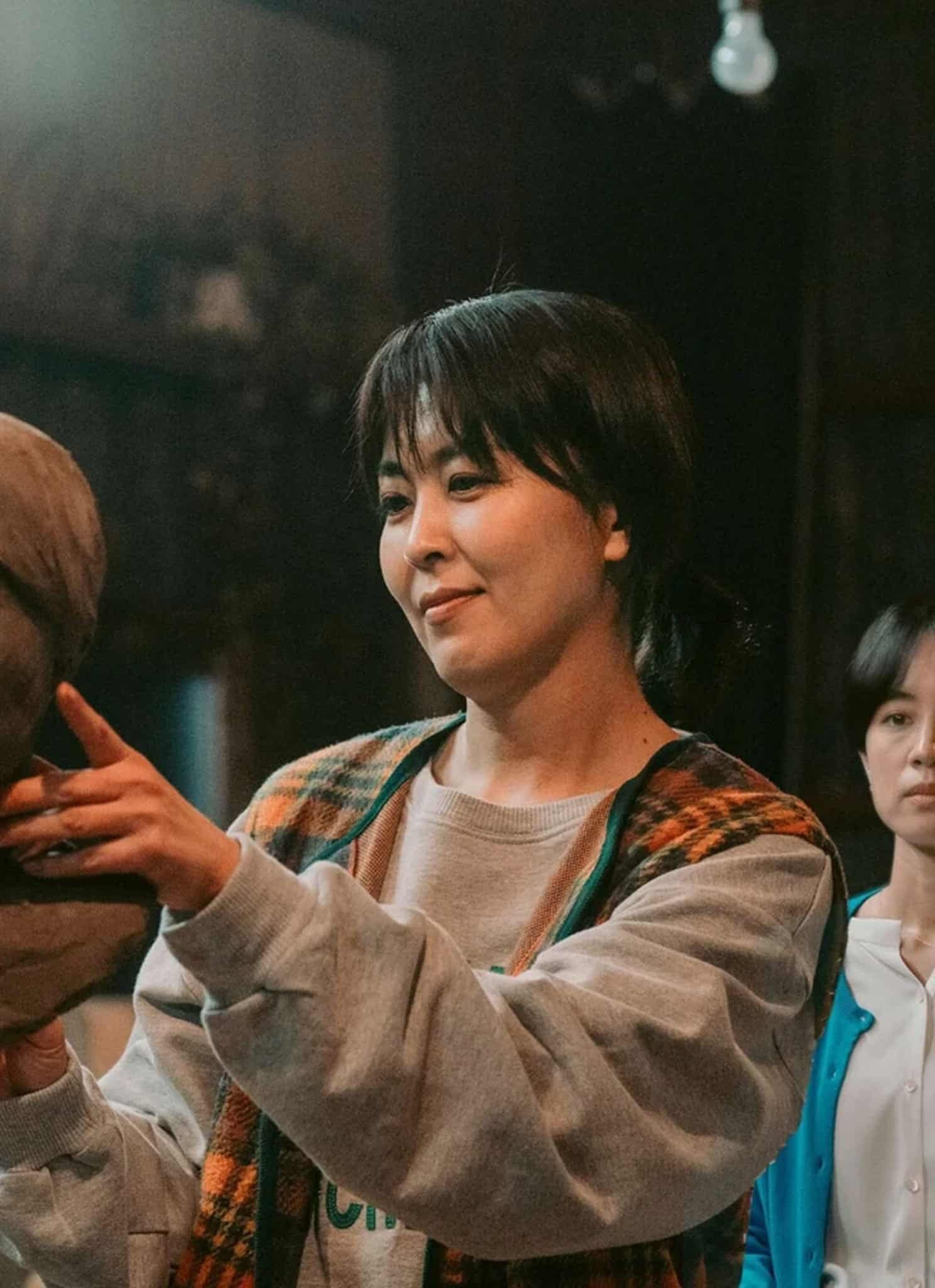 A woman in a plaid vest sculpts a clay bust, focusing intently, while another woman in a blue cardigan stands in the background watching her in a dimly lit studio.