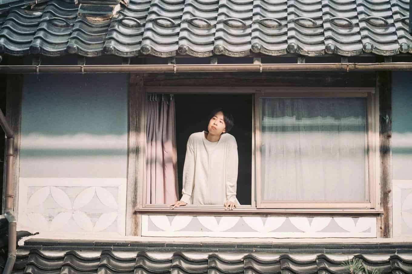 A person in a light shirt leans on the windowsill of a traditional Japanese-style house, looking up thoughtfully. The window is open, with pink curtains and patterned wall details visible.