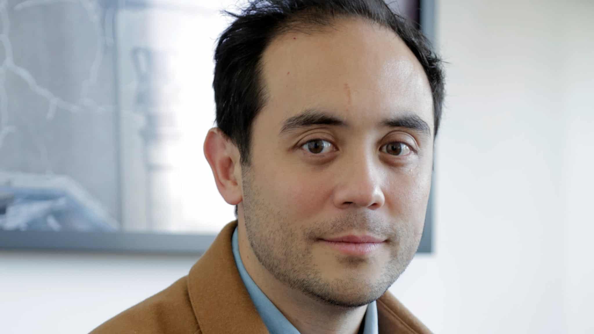 A man with short dark hair and light skin wearing a brown jacket and blue shirt looks at the camera. The background is softly blurred, showing part of a framed picture and light-colored walls.