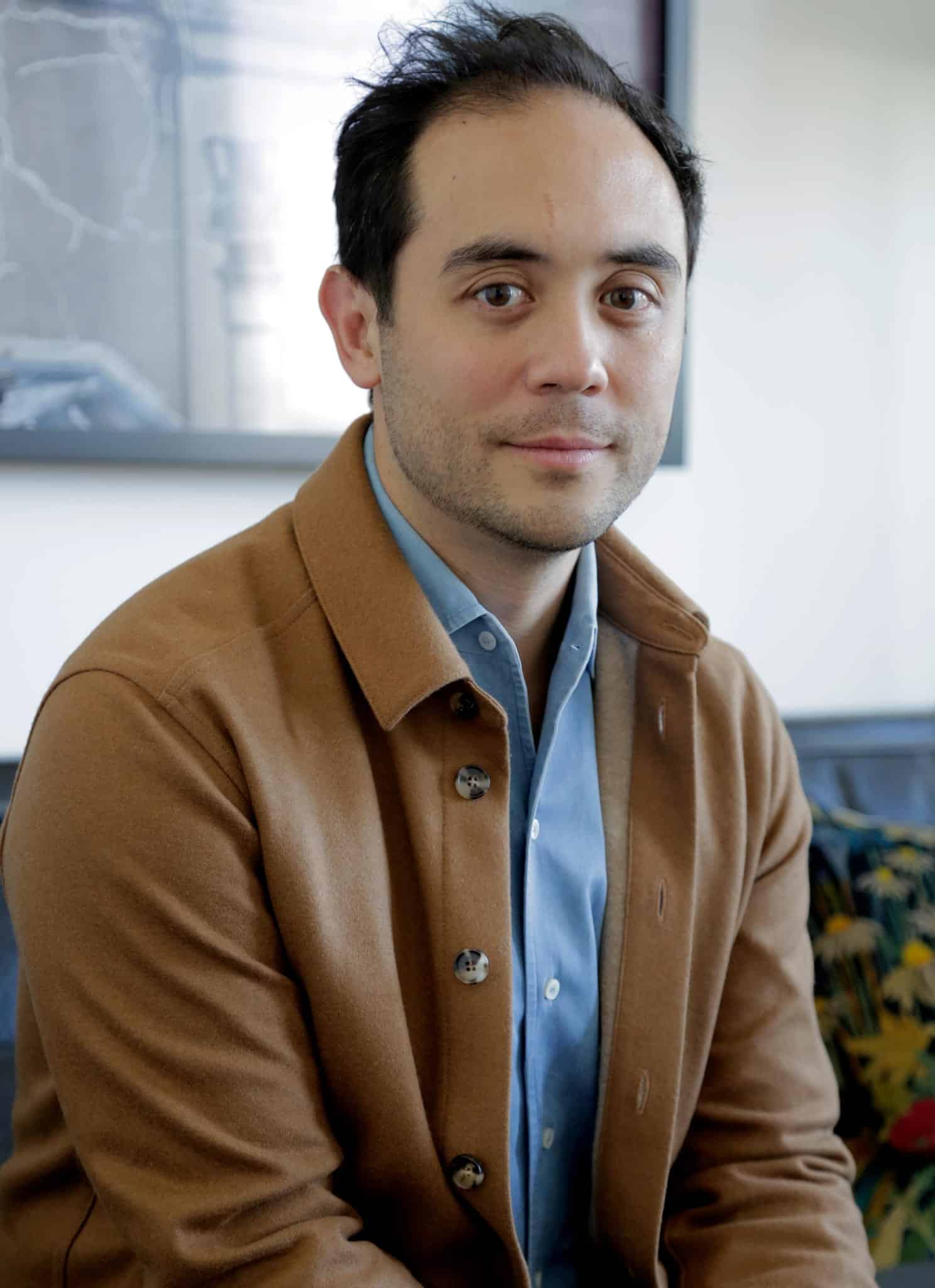 A man with short dark hair and light skin wearing a brown jacket and blue shirt looks at the camera. The background is softly blurred, showing part of a framed picture and light-colored walls.