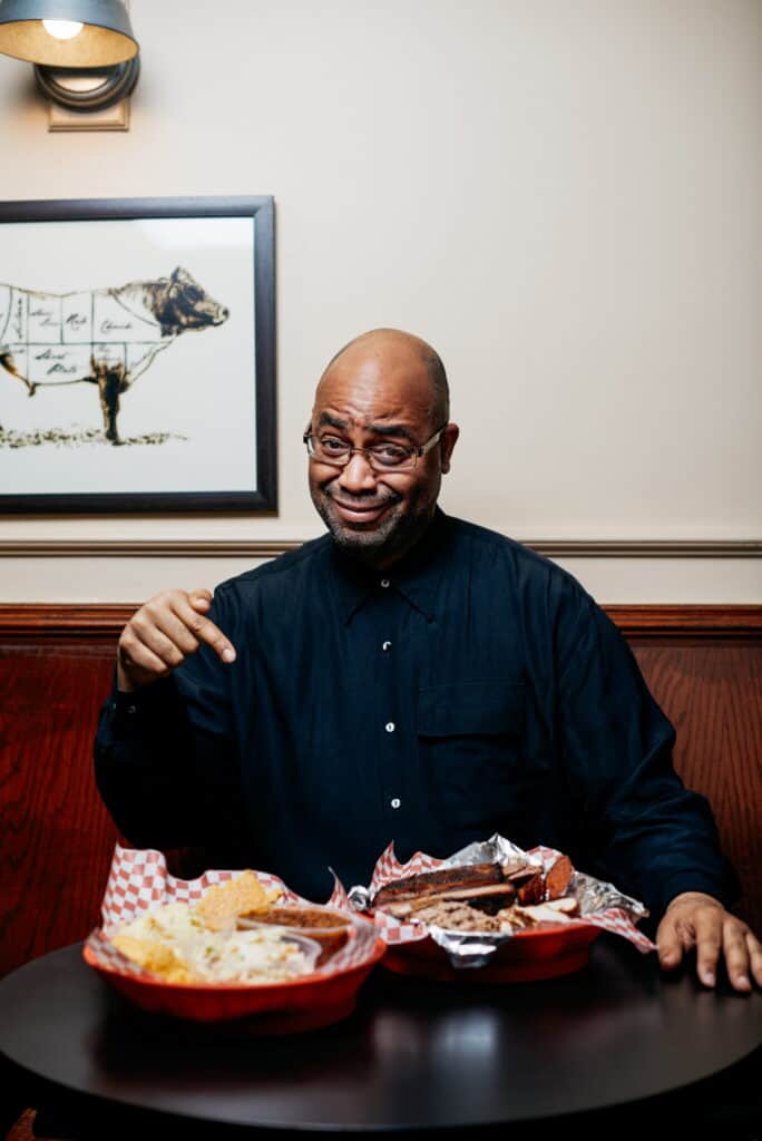 A man in glasses and a dark shirt sits at a table pointing at plates of food, including barbecue and sides, smiling at the camera. A framed drawing of a cow hangs on the wall behind him.