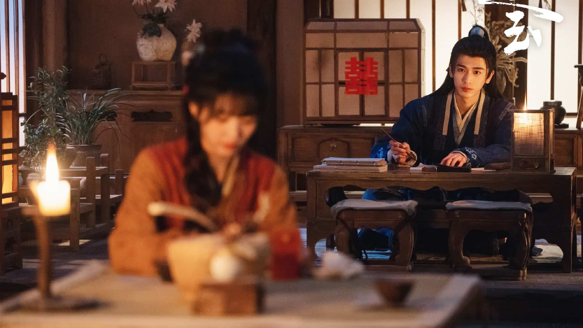 A woman in traditional Chinese attire, writes at a candle-lit table in the foreground while a man in blue traditional clothing sits at another table, looking at her in a warmly lit room with wooden decor.