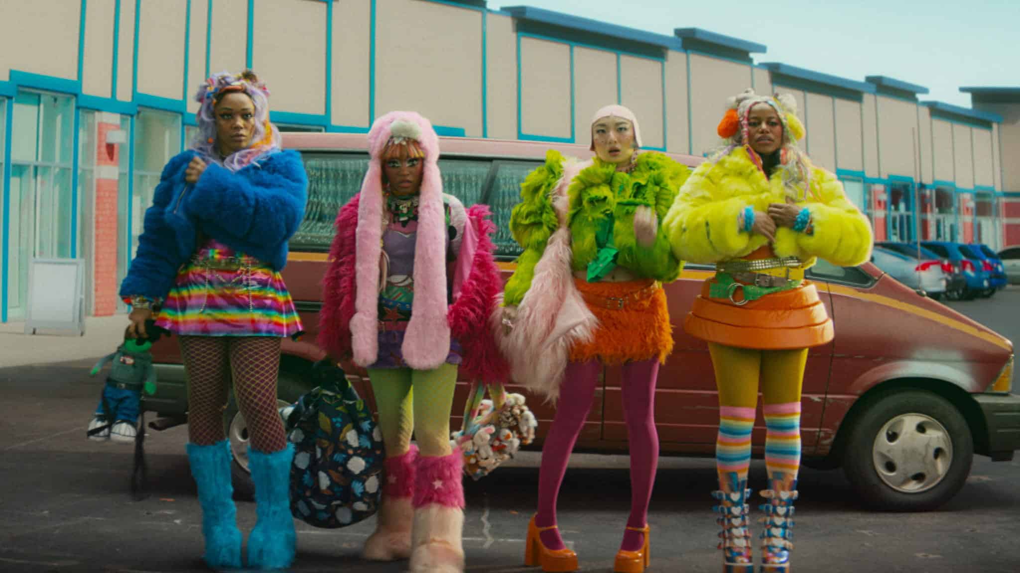 Four people in brightly colored, eccentric outfits stand in front of a red van in a parking lot, with a building and parked cars in the background. Each person wears bold patterns, fluffy textures, and vibrant accessories.