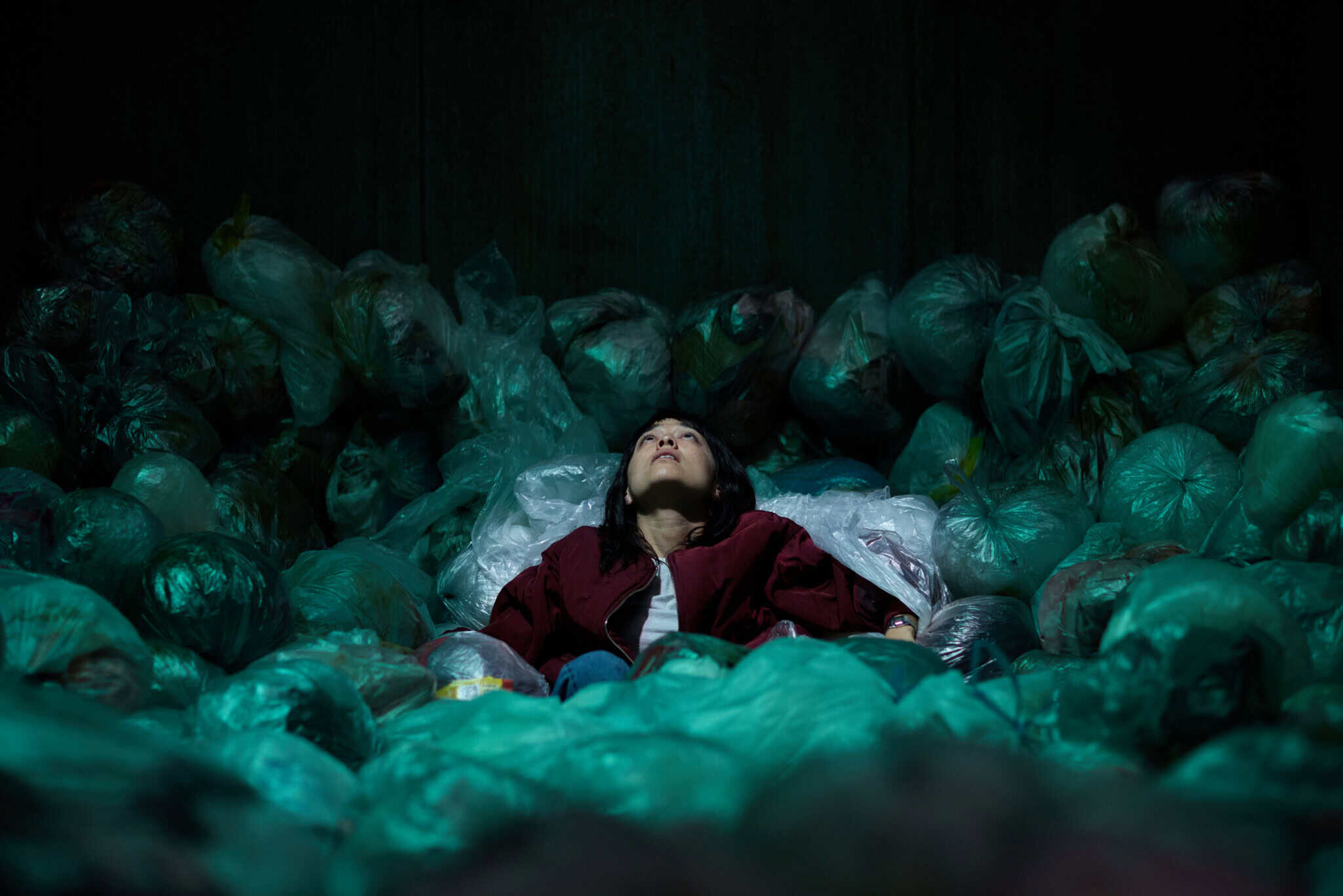 A person in a red jacket lies on their back, looking upward, surrounded by numerous green and blue plastic garbage bags in a dimly lit setting.