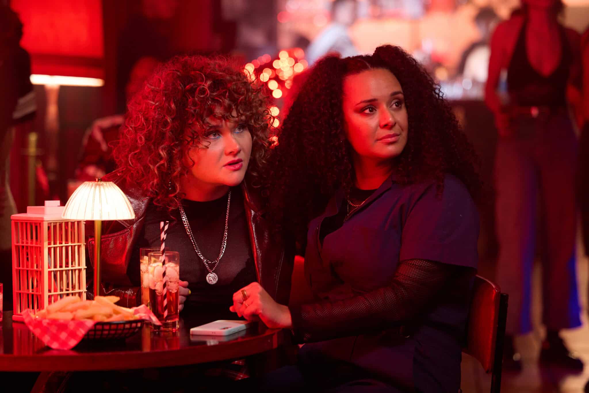 Two women with curly hair sit closely together at a dimly lit bar table with drinks and fries. They look intently in the same direction, illuminated by warm red lighting and small glowing lamps, with other people blurred in the background.