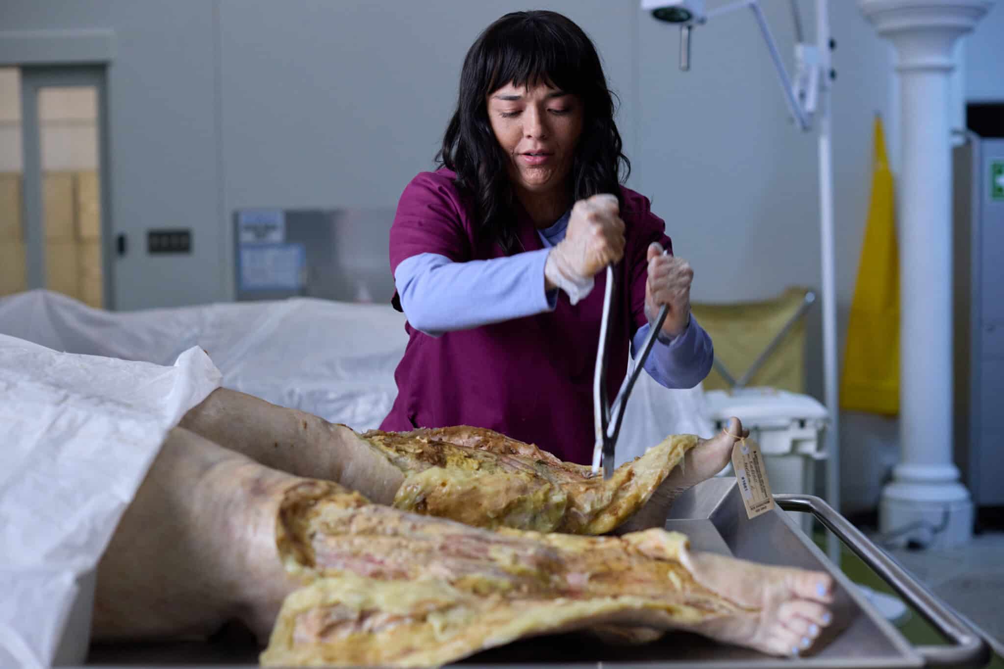 A person in scrubs examines or works on a cadaver on a table in a medical or laboratory setting, handling an instrument over the exposed lower body.