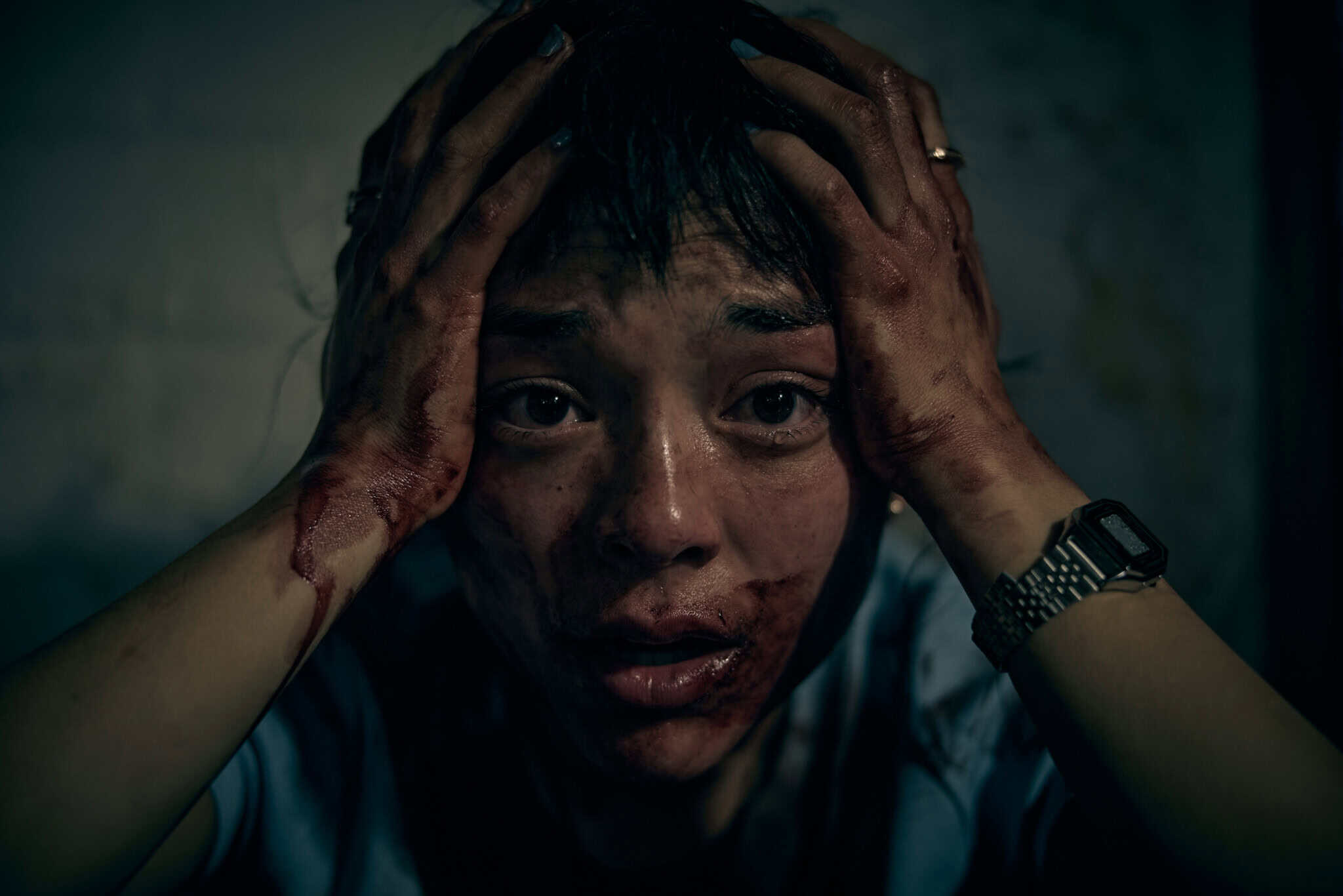 A person with blood on their face and hands sits with their head in their hands, looking distressed. The background is dimly lit, creating a dramatic and intense atmosphere.