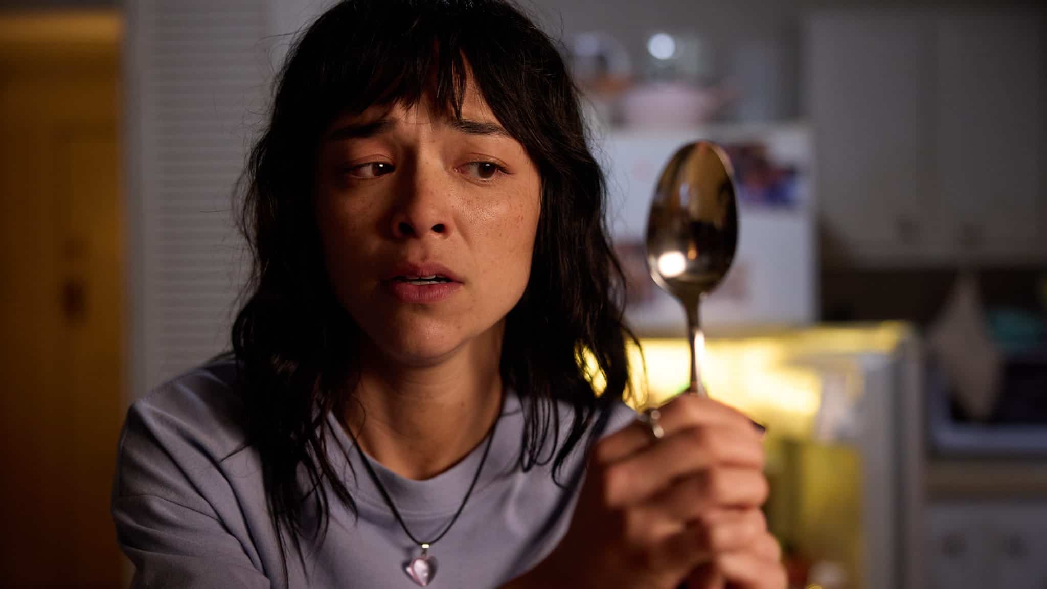 A woman with dark hair and a necklace holds up a large spoon and looks concerned or worried. She is indoors, with soft lighting and a kitchen visible in the background.