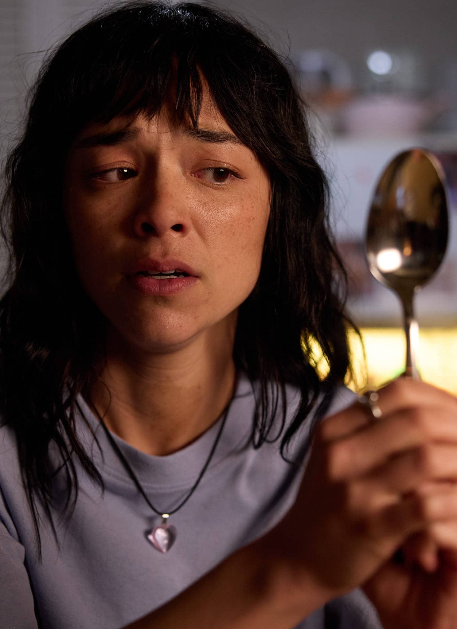A woman with dark hair and a necklace holds up a large spoon and looks concerned or worried. She is indoors, with soft lighting and a kitchen visible in the background.