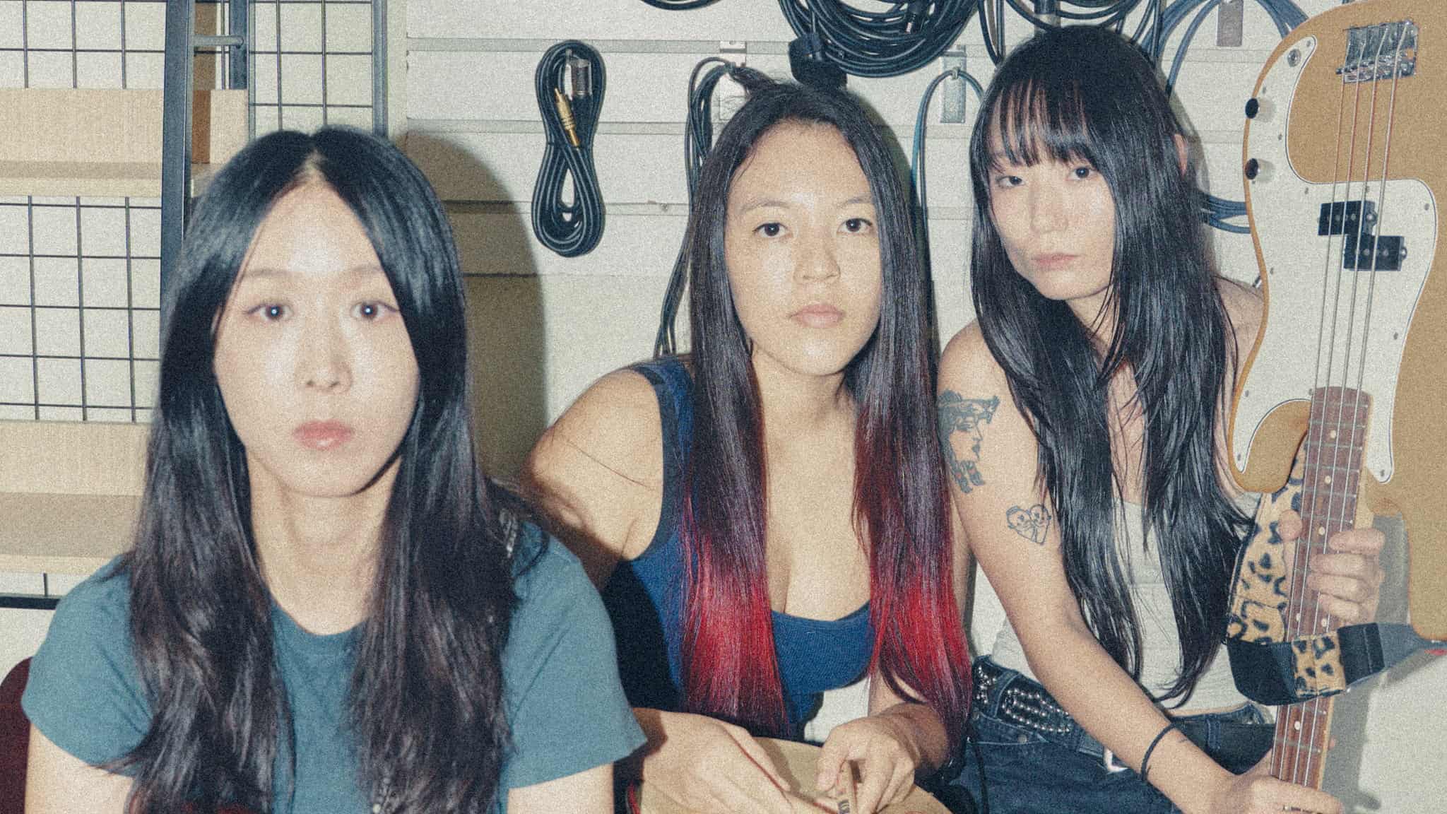 Three young women with long dark hair sit indoors surrounded by music equipment and cables; one holds an electric bass guitar. All three look directly at the camera with neutral expressions.