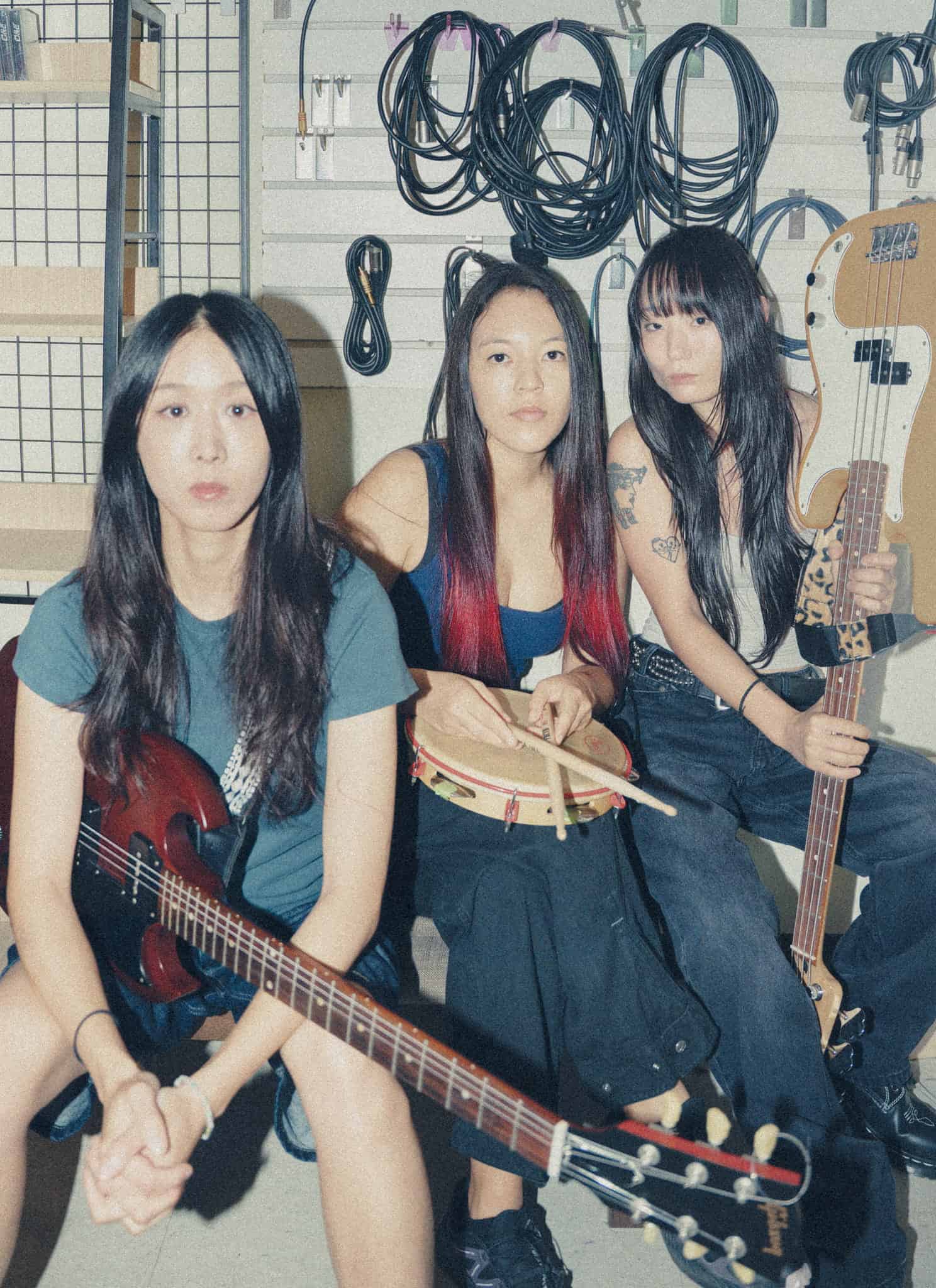 Three young women with long dark hair sit indoors surrounded by music equipment and cables; one holds an electric bass guitar. All three look directly at the camera with neutral expressions.